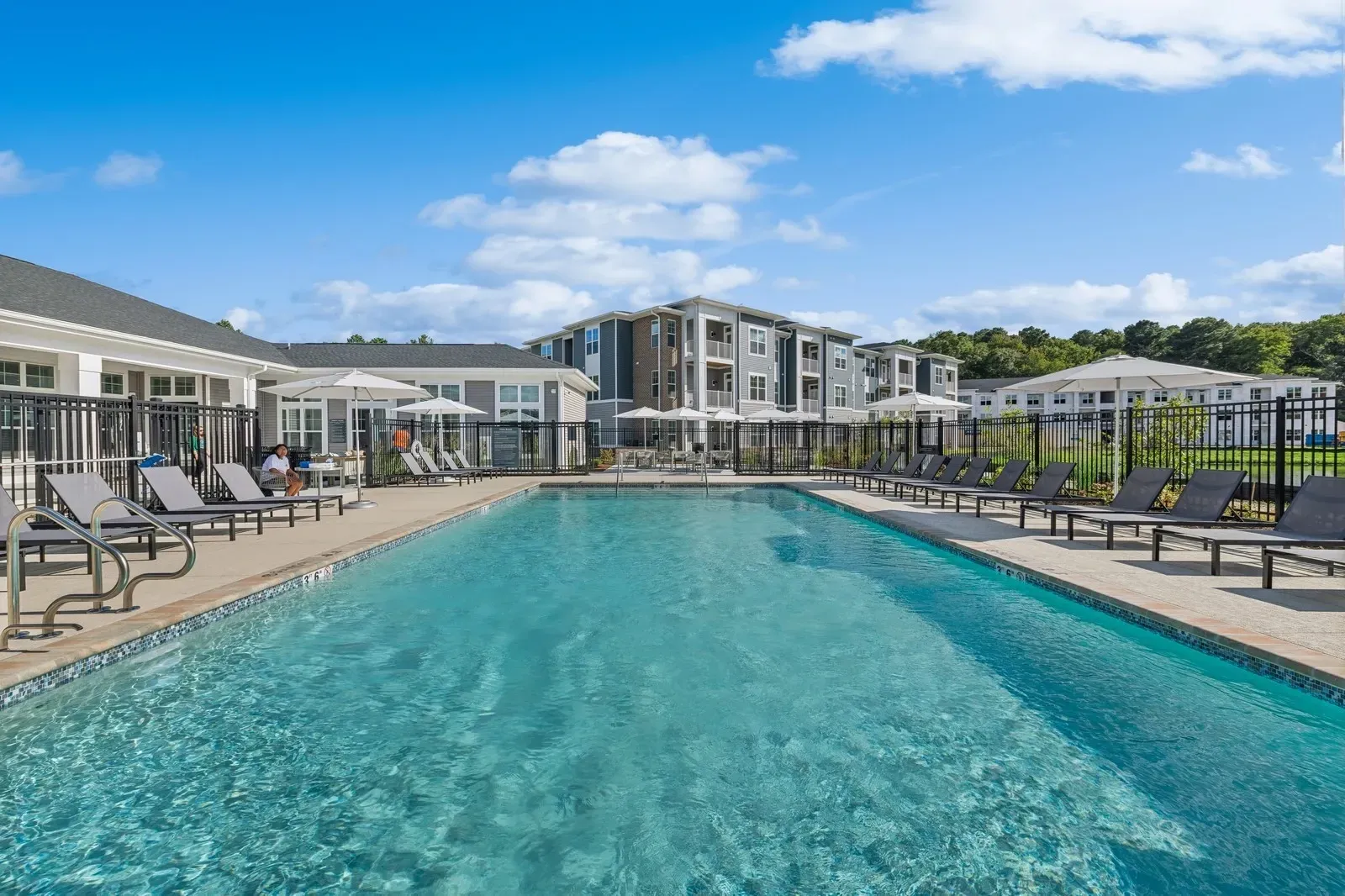Outdoor communal pool area with lounge chairs, umbrellas, and apartment buildings in the background.