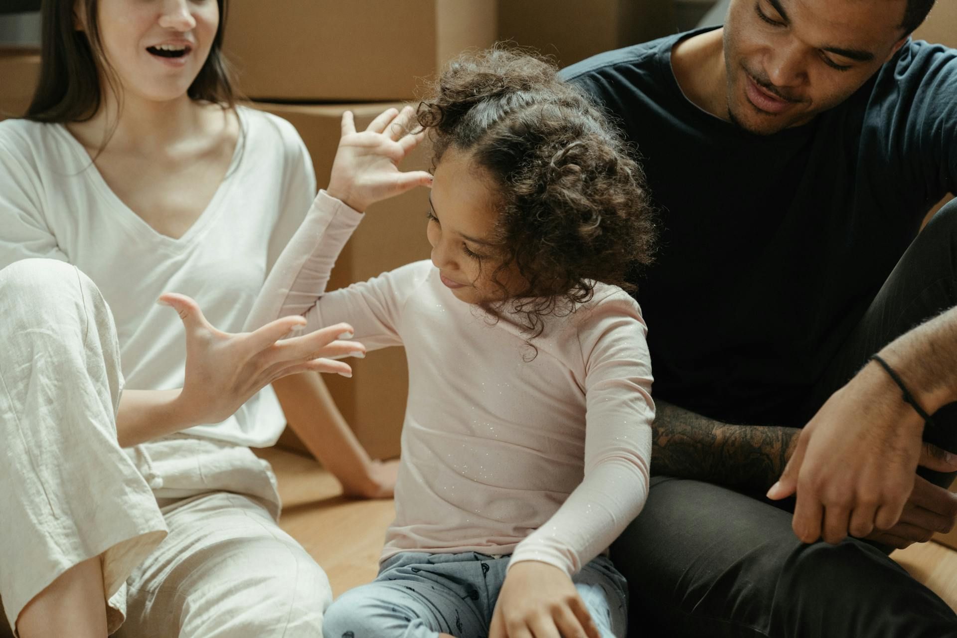 A family sitting on a floor near moving boxes, with a young child gesturing while talking to their parents.