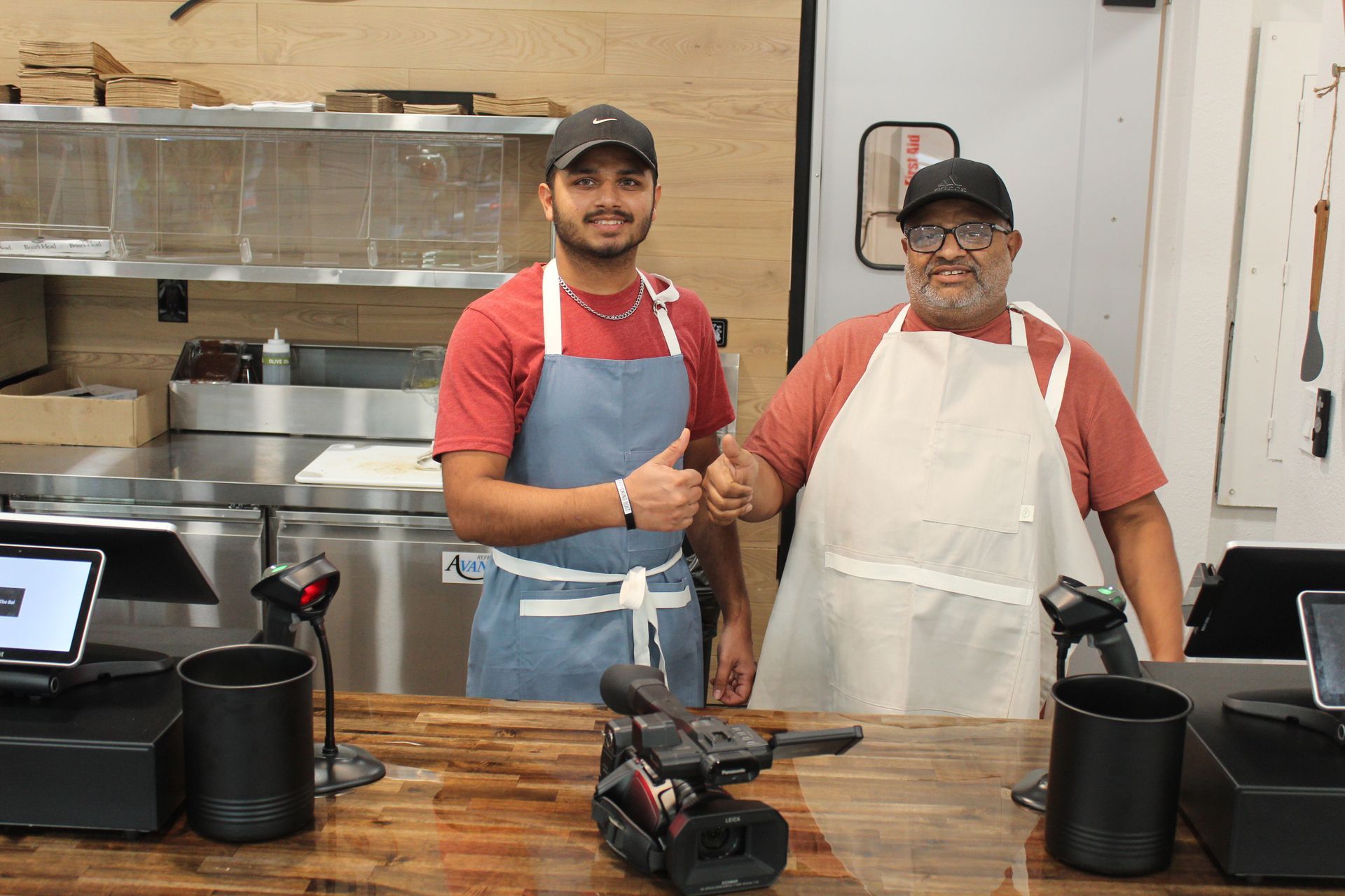 Two men are standing next to each other in a kitchen giving a thumbs up.