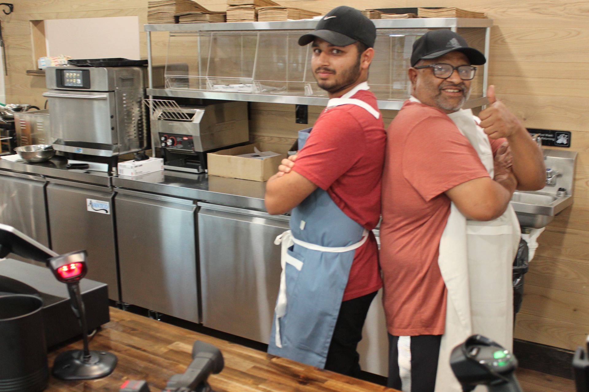 Two men are standing back to back in a kitchen giving a thumbs up.
