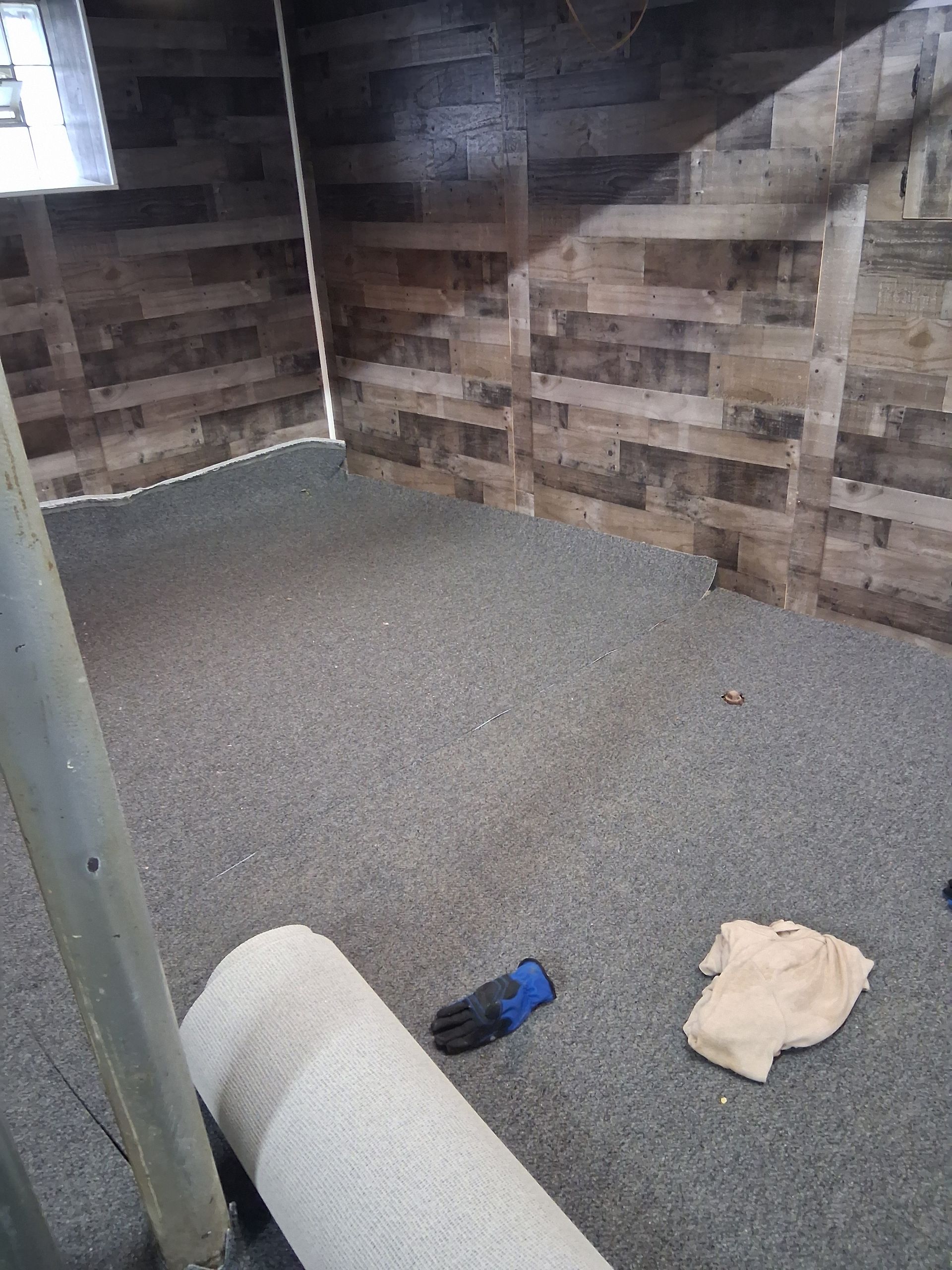 A room with wood-paneled walls and gray flooring; a rolled-up rug, glove, and cloth on the floor.