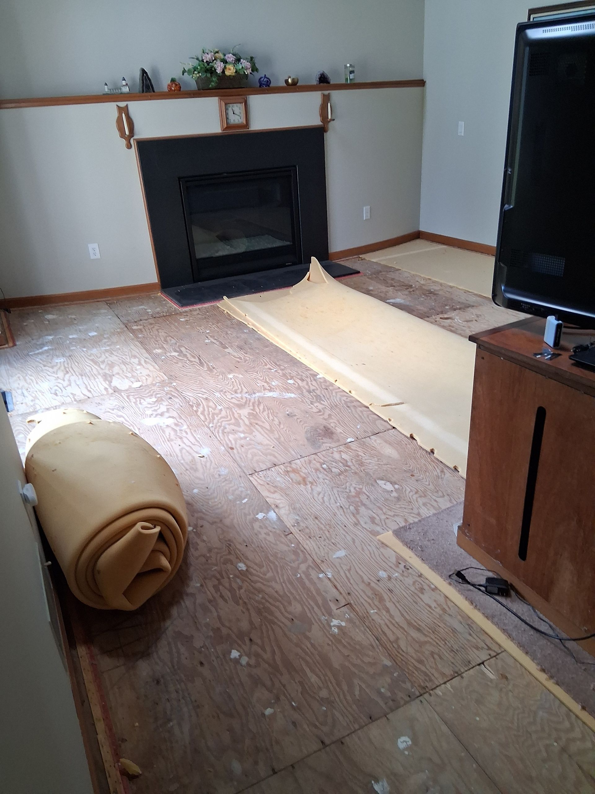 Living room with old flooring removed, roll of padding, fireplace, and TV stand.