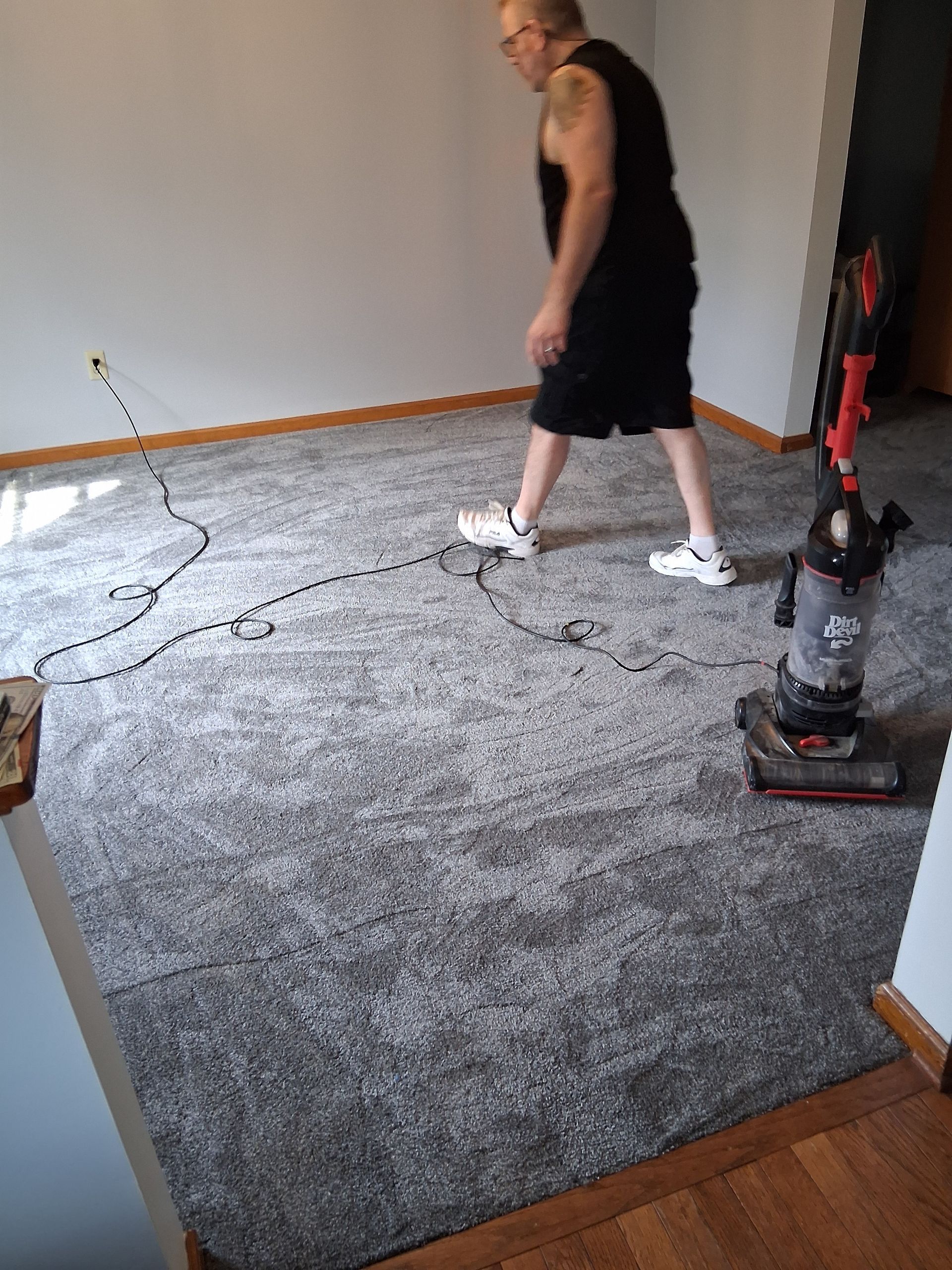 Man vacuuming a gray carpet in an empty room with white walls and wooden trim.