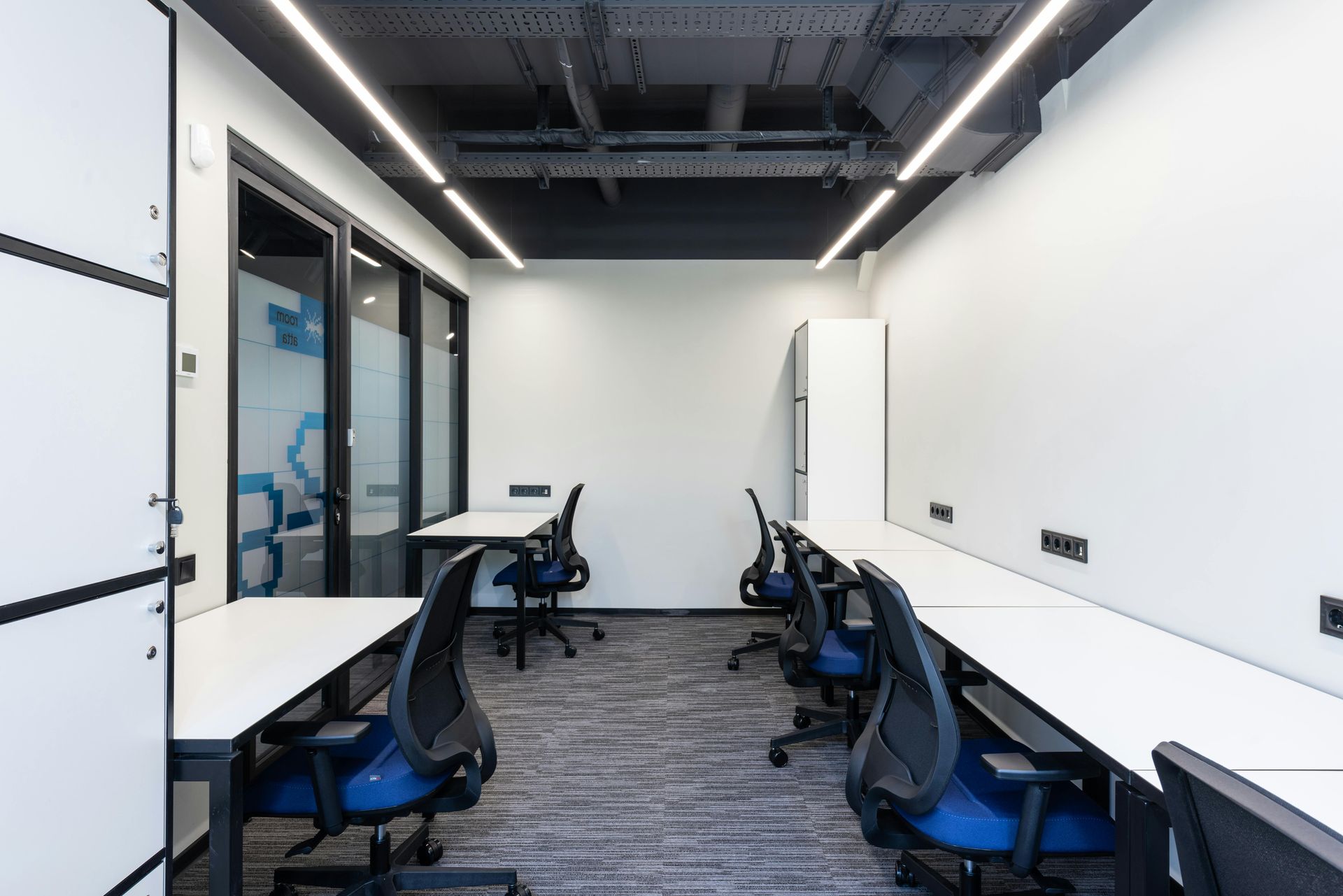 Office space with desks, chairs, and a glass-walled room. White walls, blue chairs, and gray carpet.