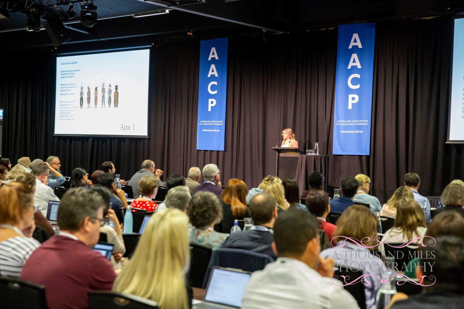 Audience at conference listening to a presenter at the podium under AACP banners.