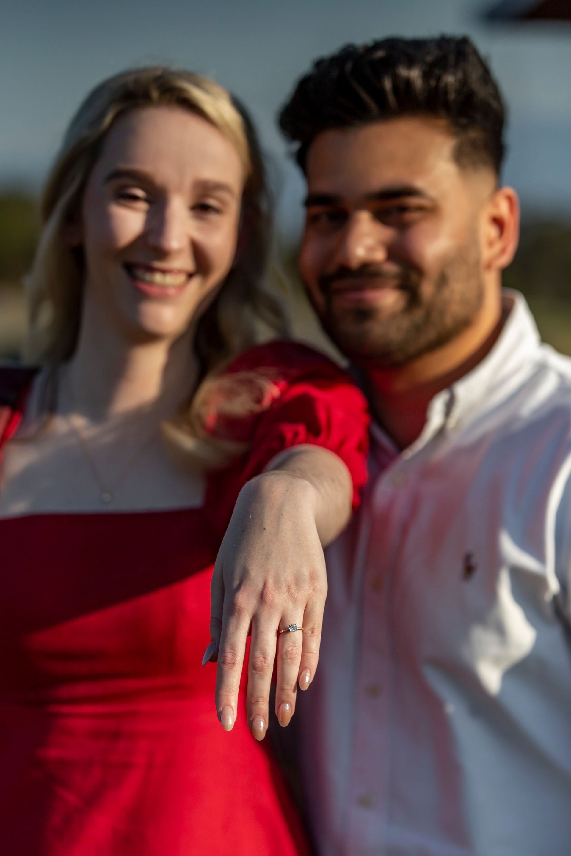 Engaged couple: woman in red dress showing ring, man in white shirt smiles outdoors.