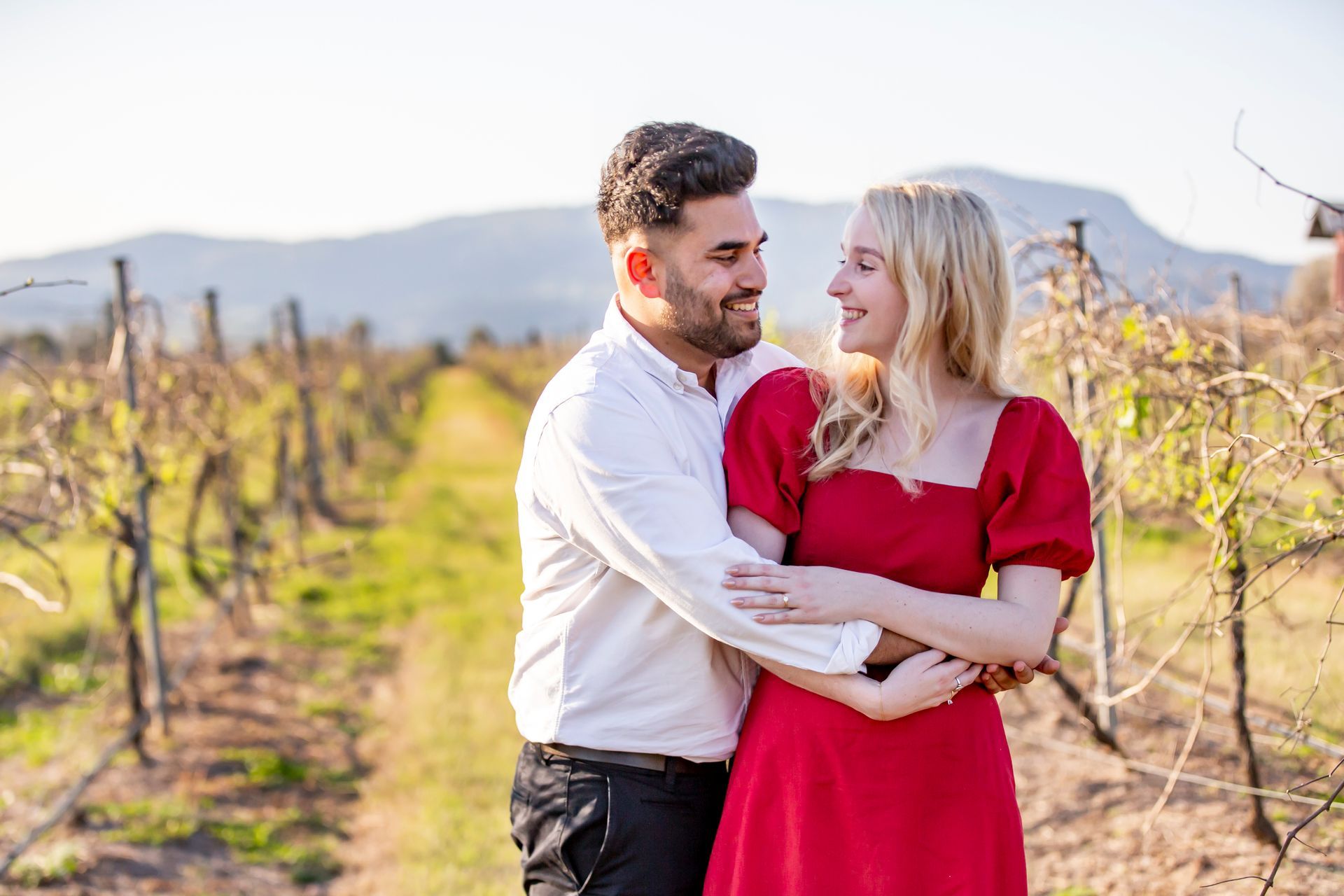 Couple embracing in vineyard, man in white shirt, woman in red dress, smiling, mountain backdrop.