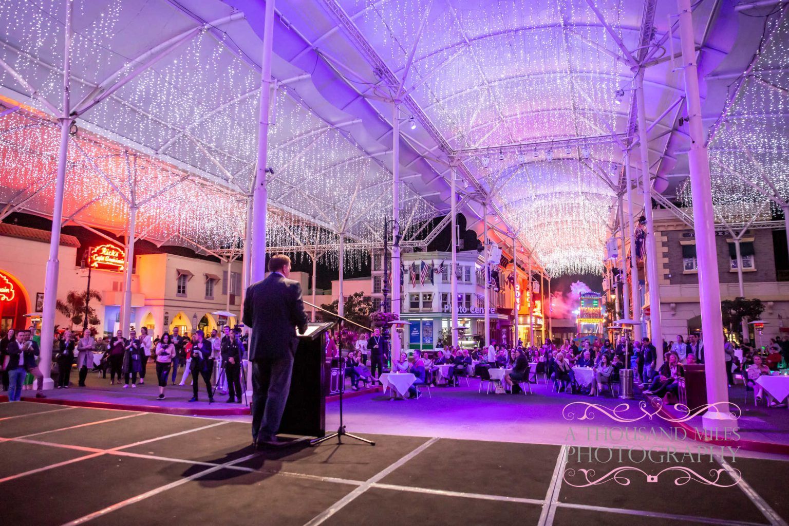 Man speaking at podium in front of a crowd at night under purple lights.