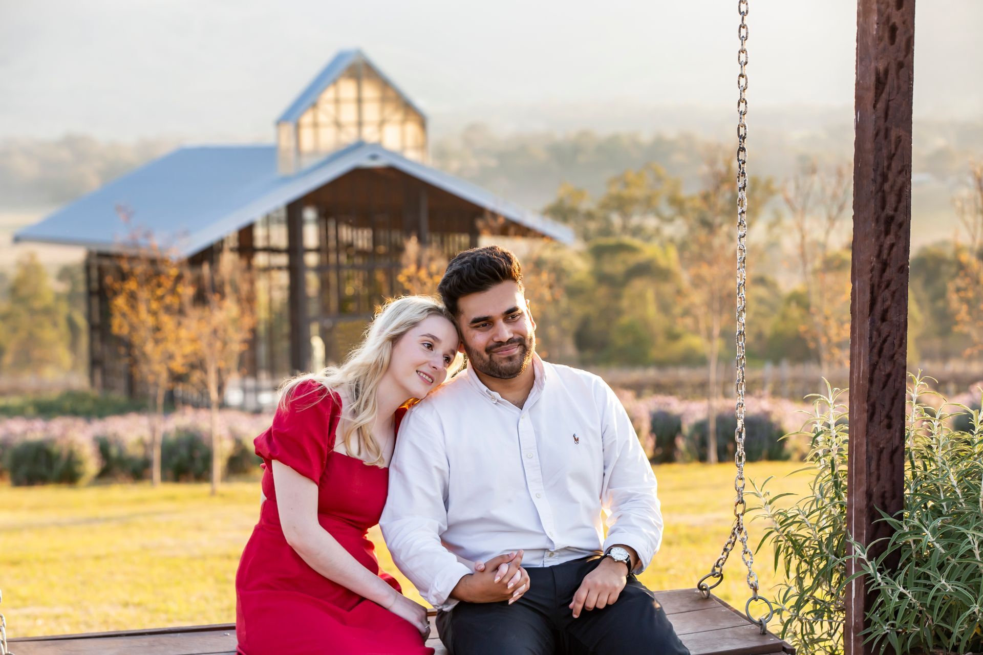 Couple on a swing, woman in red dress leans on man in white shirt, with a building in the background.