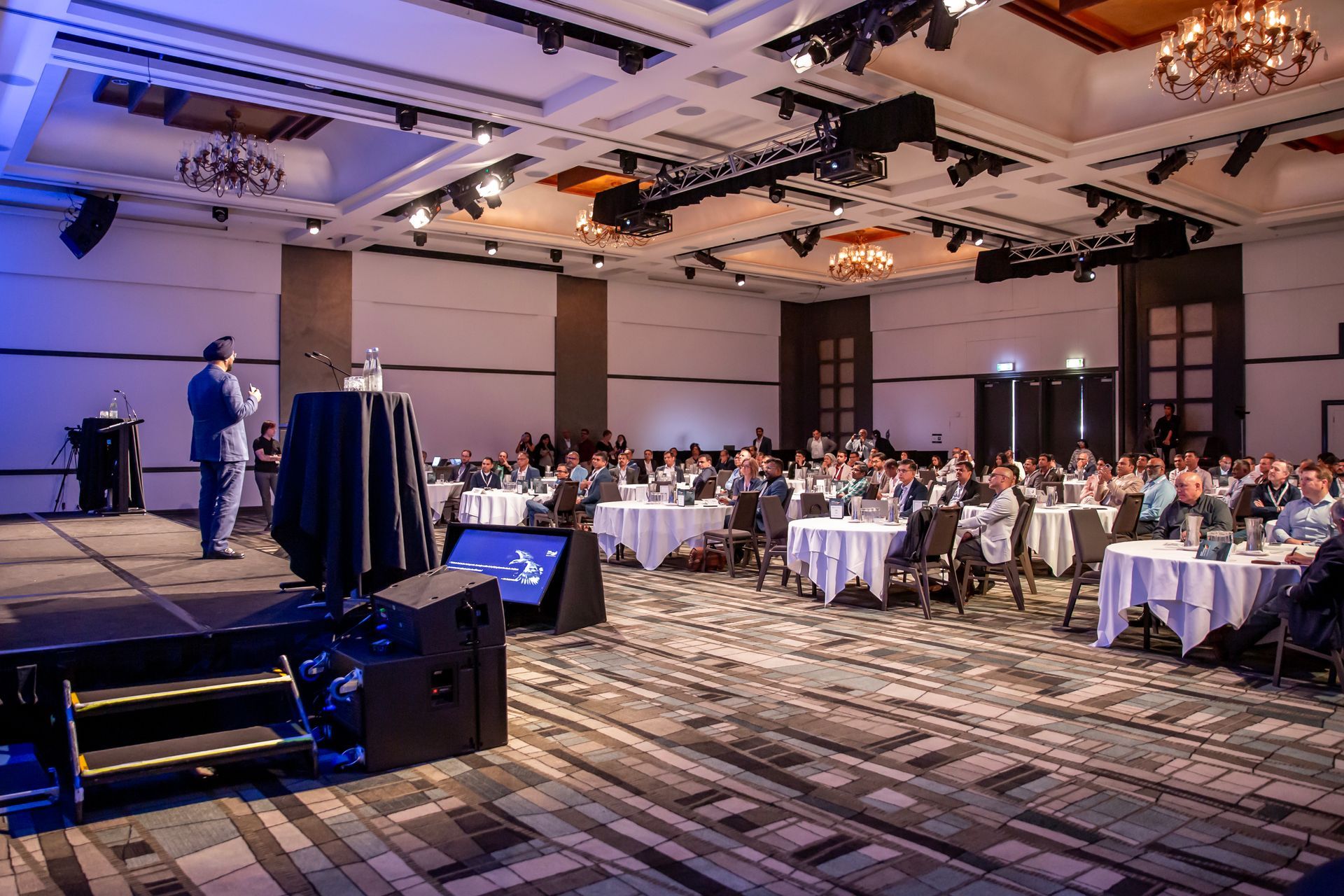 Man speaking at conference; people seated at tables in ballroom listening.