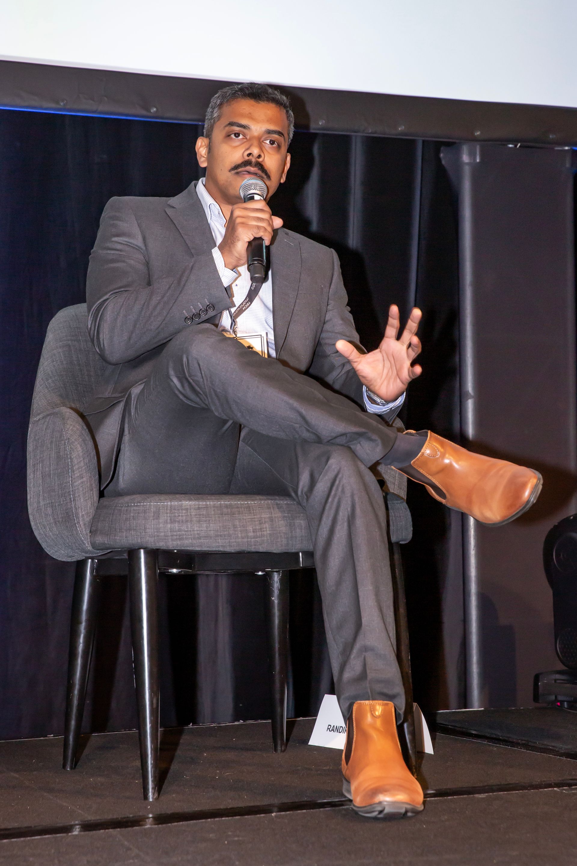 Man with mustache in gray suit and brown boots speaks into a microphone, seated on stage.