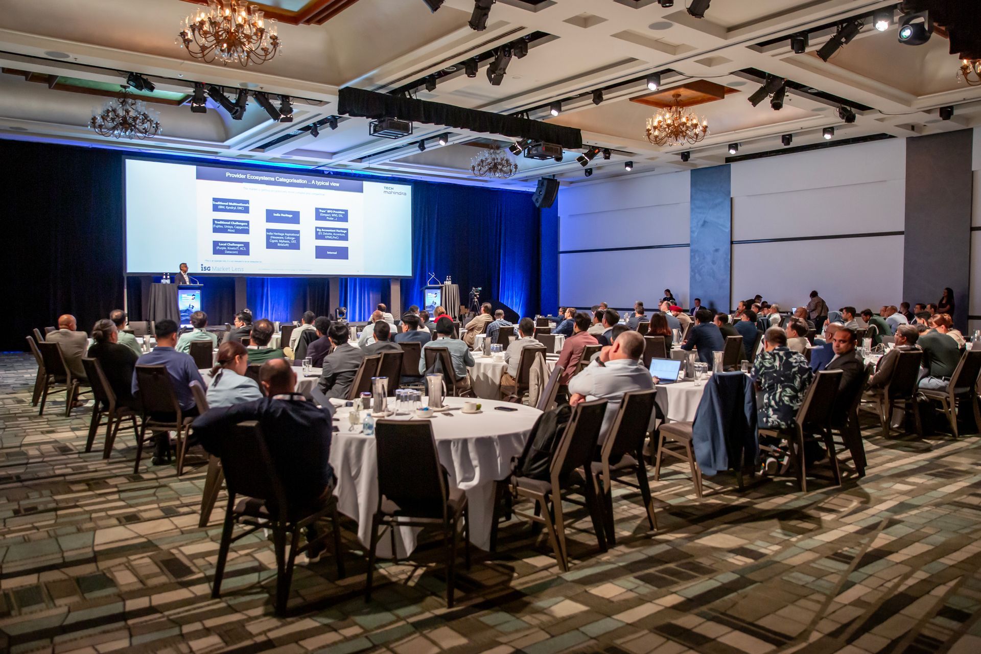 Large conference room with attendees seated at tables, listening to a presentation on a screen.