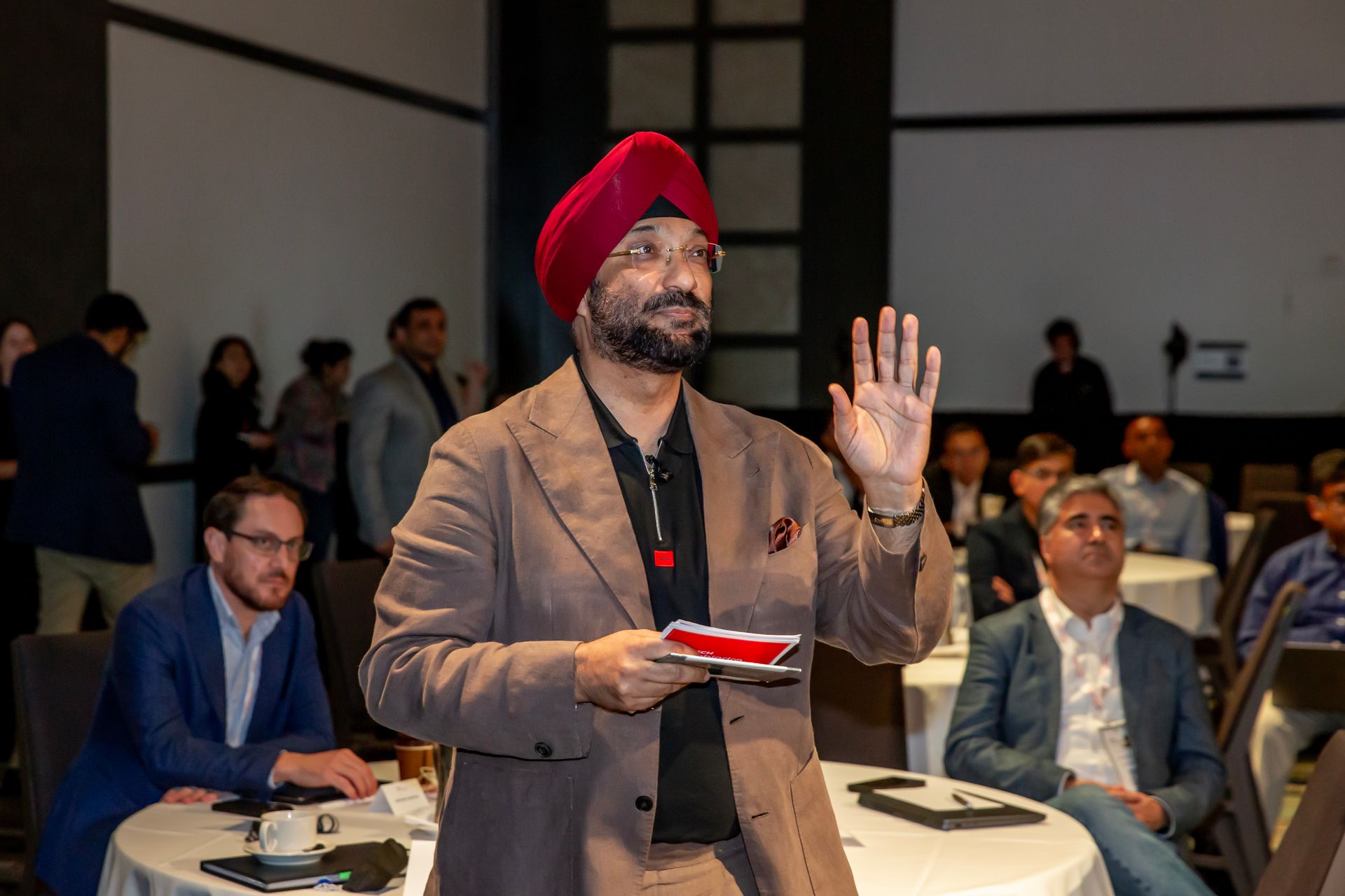 Man in turban gestures while speaking at event, brown suit, raising hand.