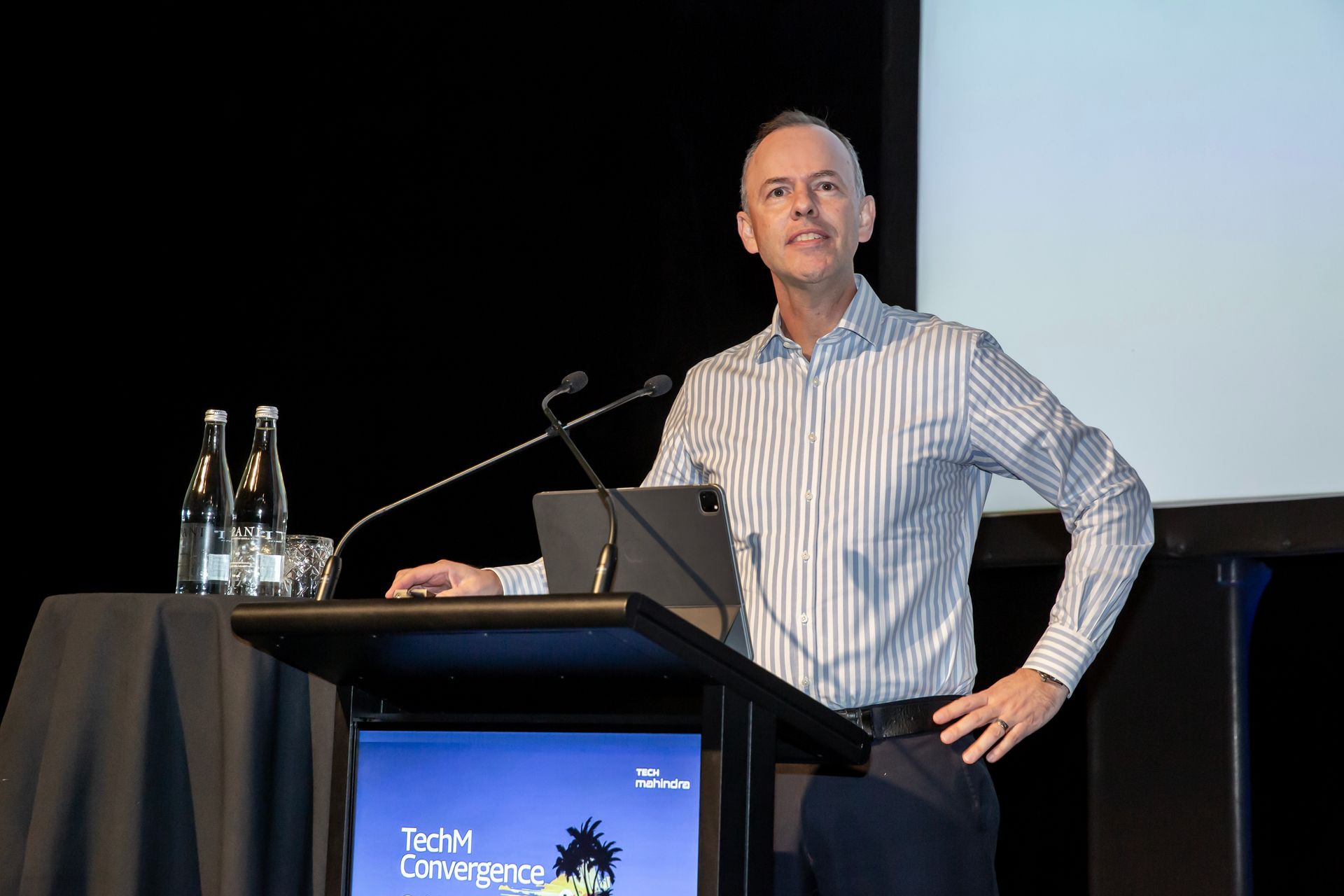 Man in striped shirt speaking at a podium, gesturing with his hand. Stage with a screen and water bottles.