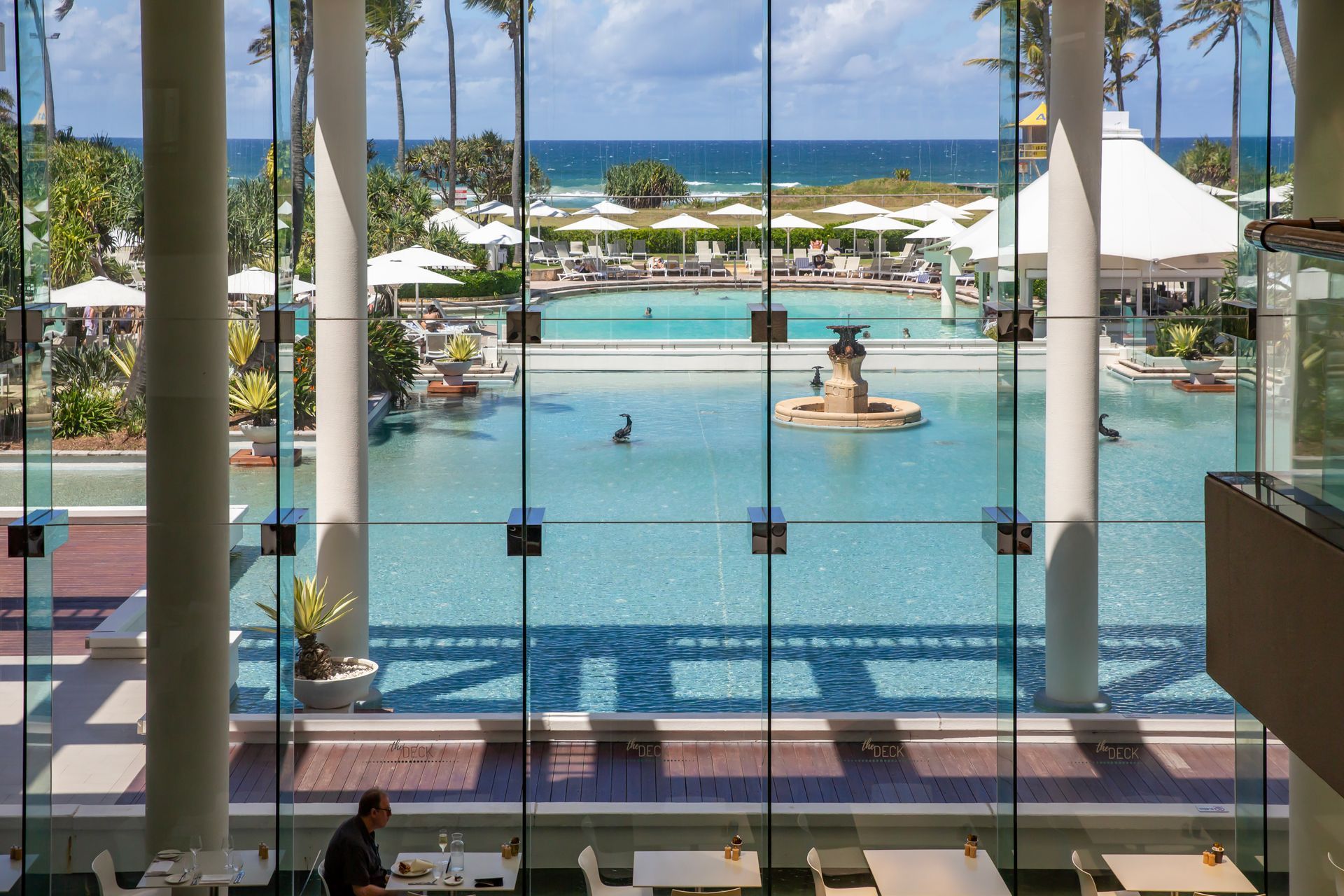 Large glass windows frame a pool with ocean view. People swim and lounge; white umbrellas provide shade.