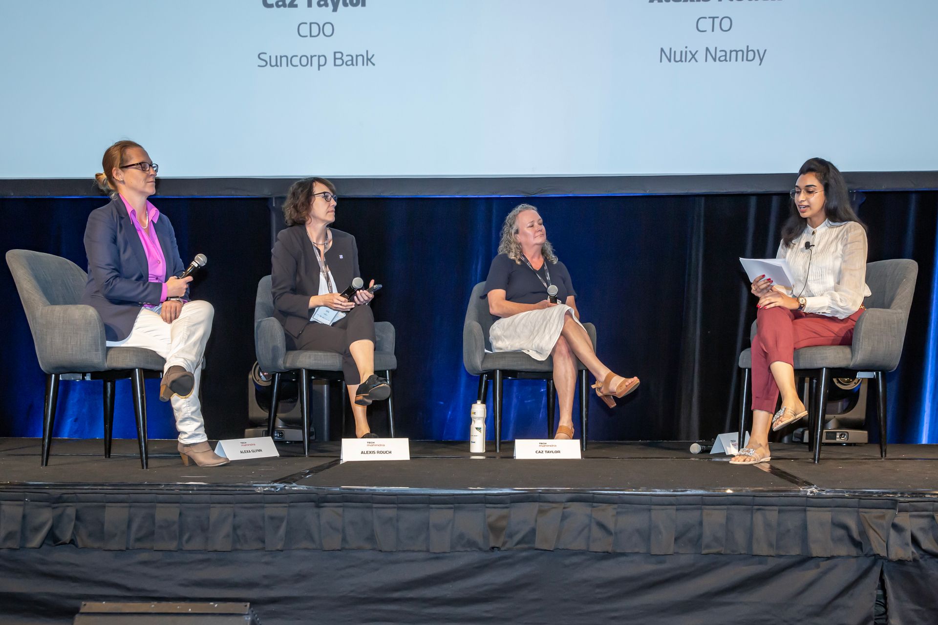 Four women on stage at a conference, seated, discussing topics. They're facing the audience, wearing business casual attire.