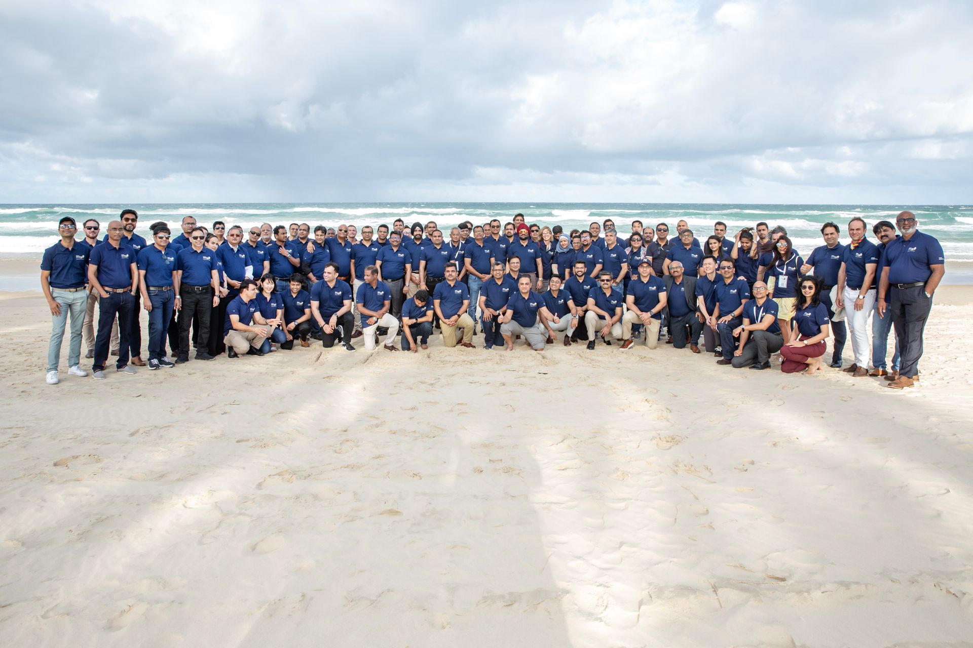Large group of people in blue shirts on a beach, posing for a photo under a cloudy sky.