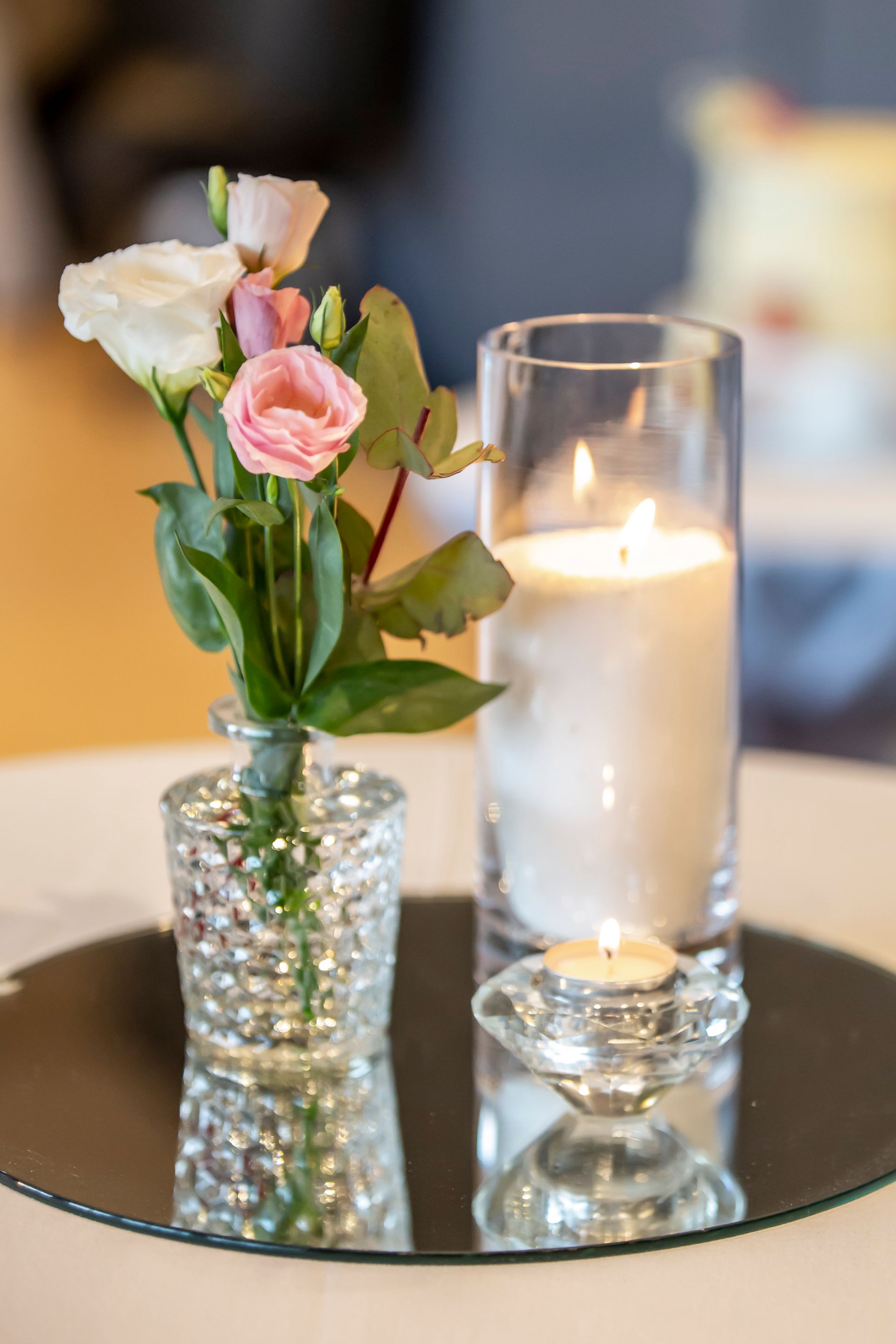 Flowers in a small vase, lit candles in glass holders, all on a mirrored surface. Photography by Andy Williams
