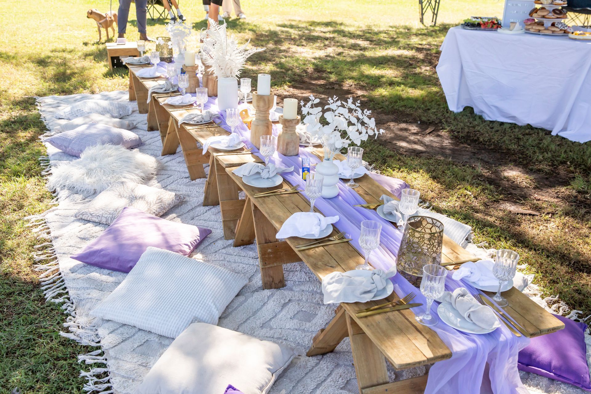 Picnic setup on grass: long wooden table with purple accents, pillows, and white floral decorations.