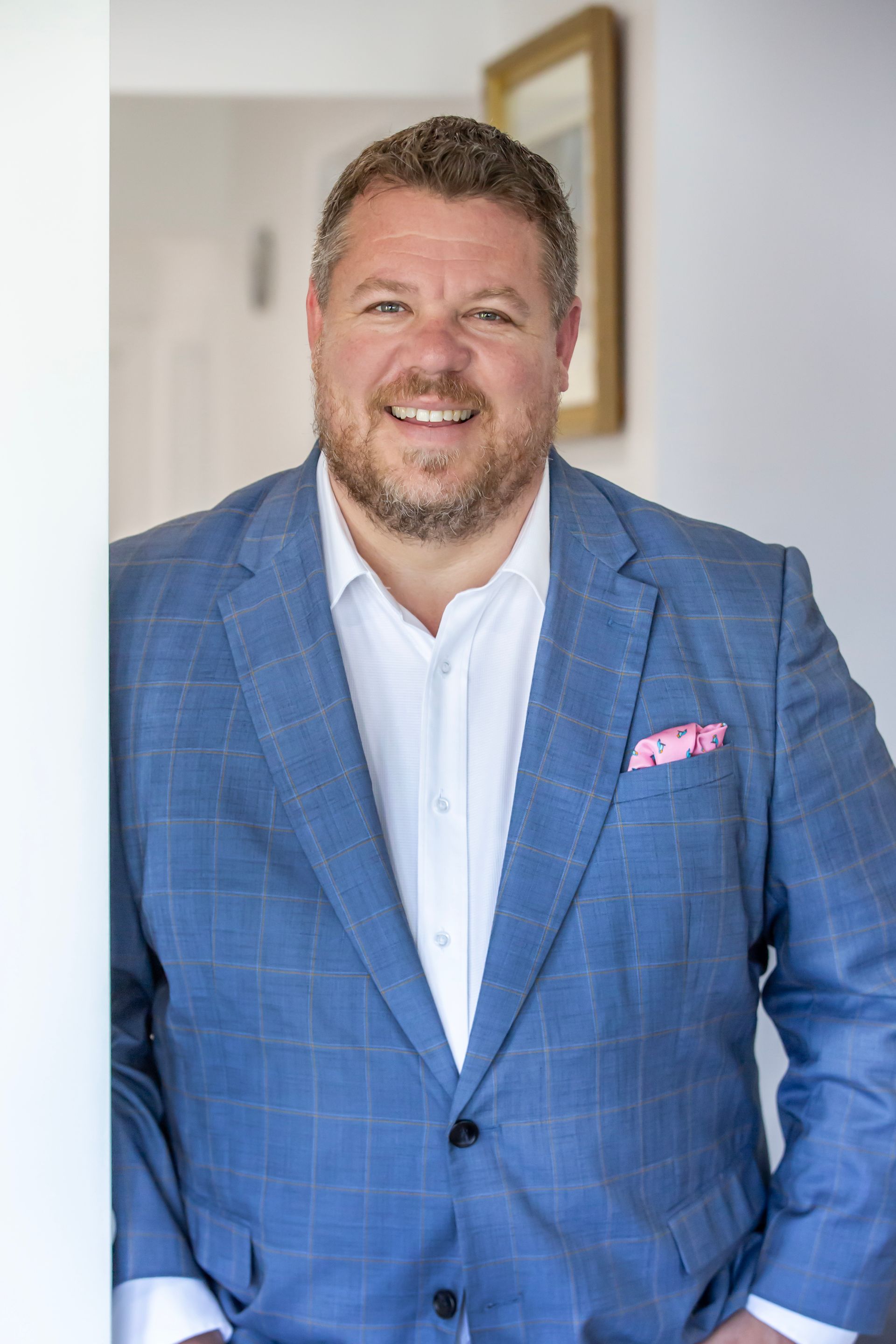 Man in a blue blazer with a pink pocket square smiles at the camera in a bright hallway.