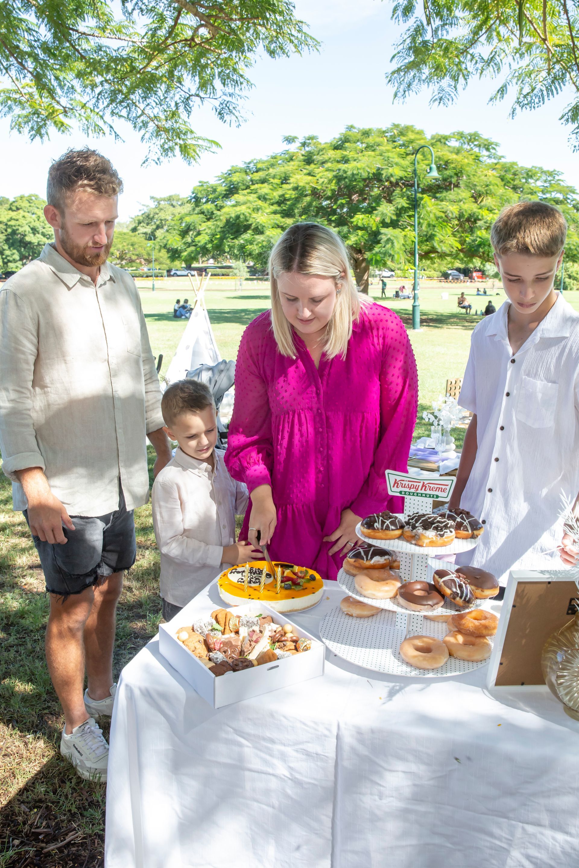 Family at a picnic table: woman cuts cake, boys watch, man looks on, donuts, bright pink dress, park setting.