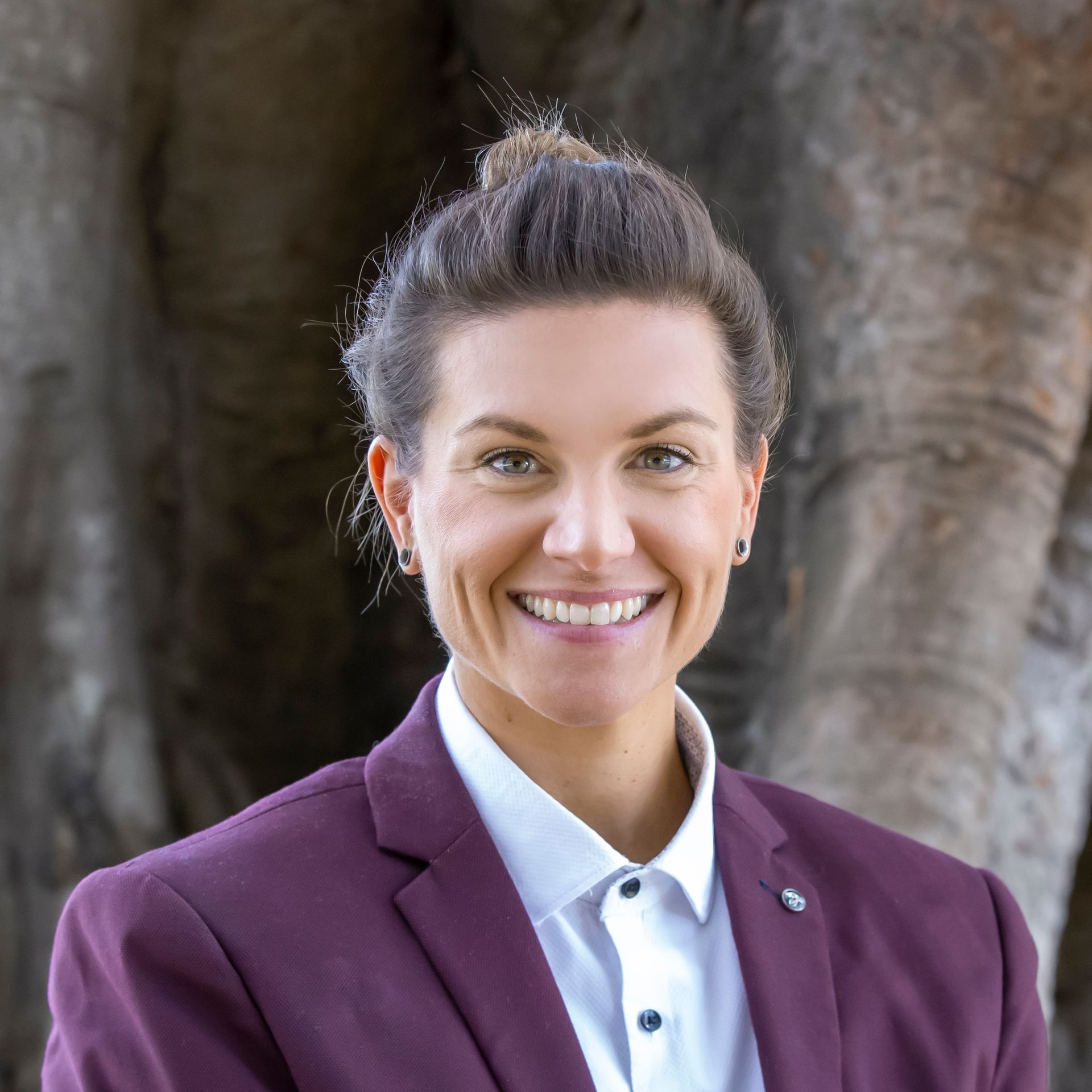 Woman in maroon blazer smiles, standing in front of a tree.