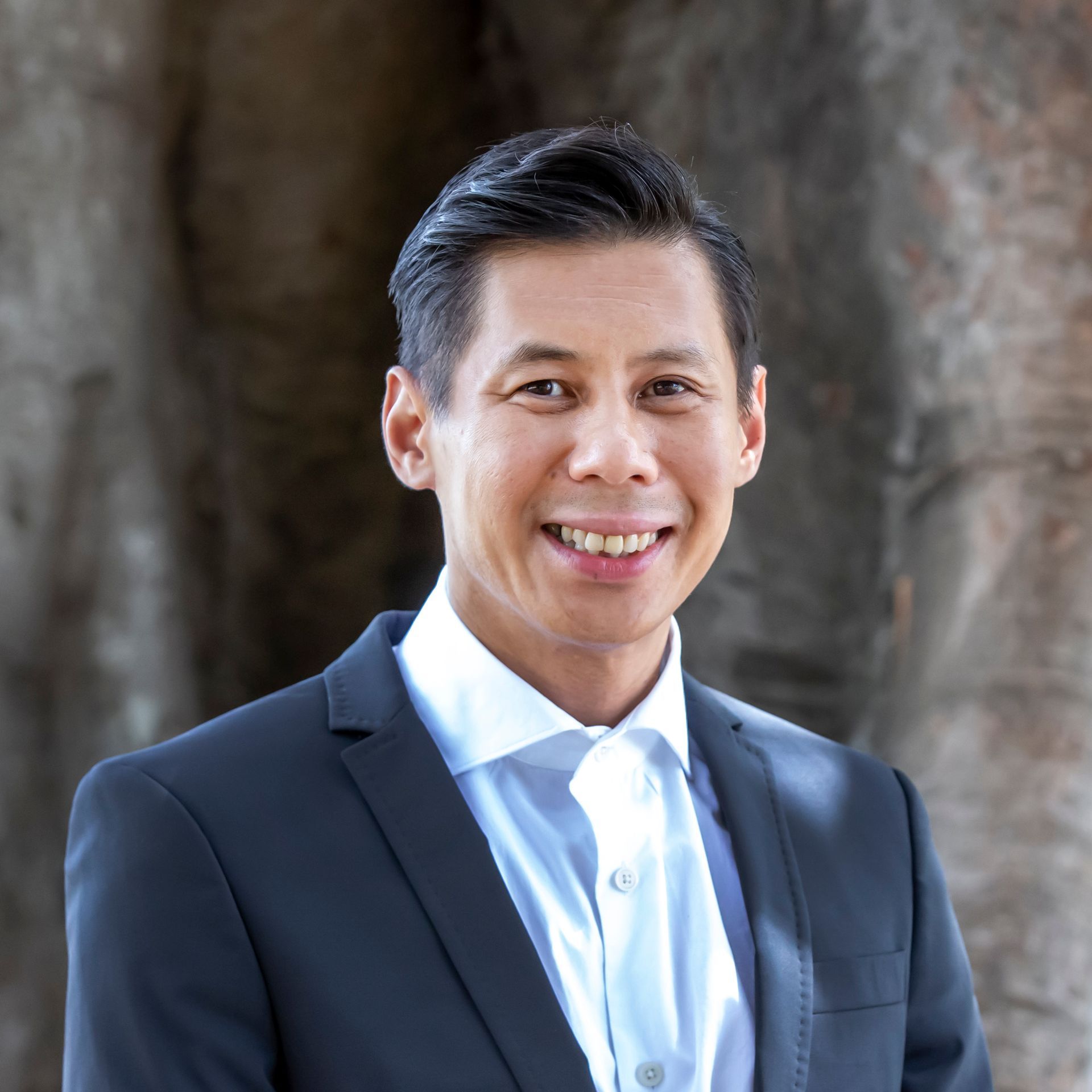 Man in dark suit, smiling, standing in front of a stone structure.