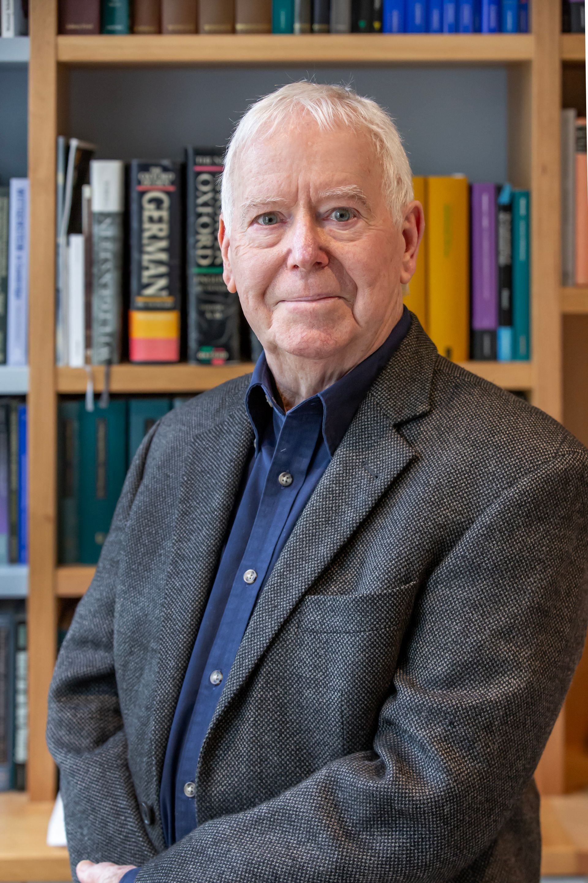 Older man with white hair and glasses wearing a dark blazer and blue shirt in front of a bookshelf.