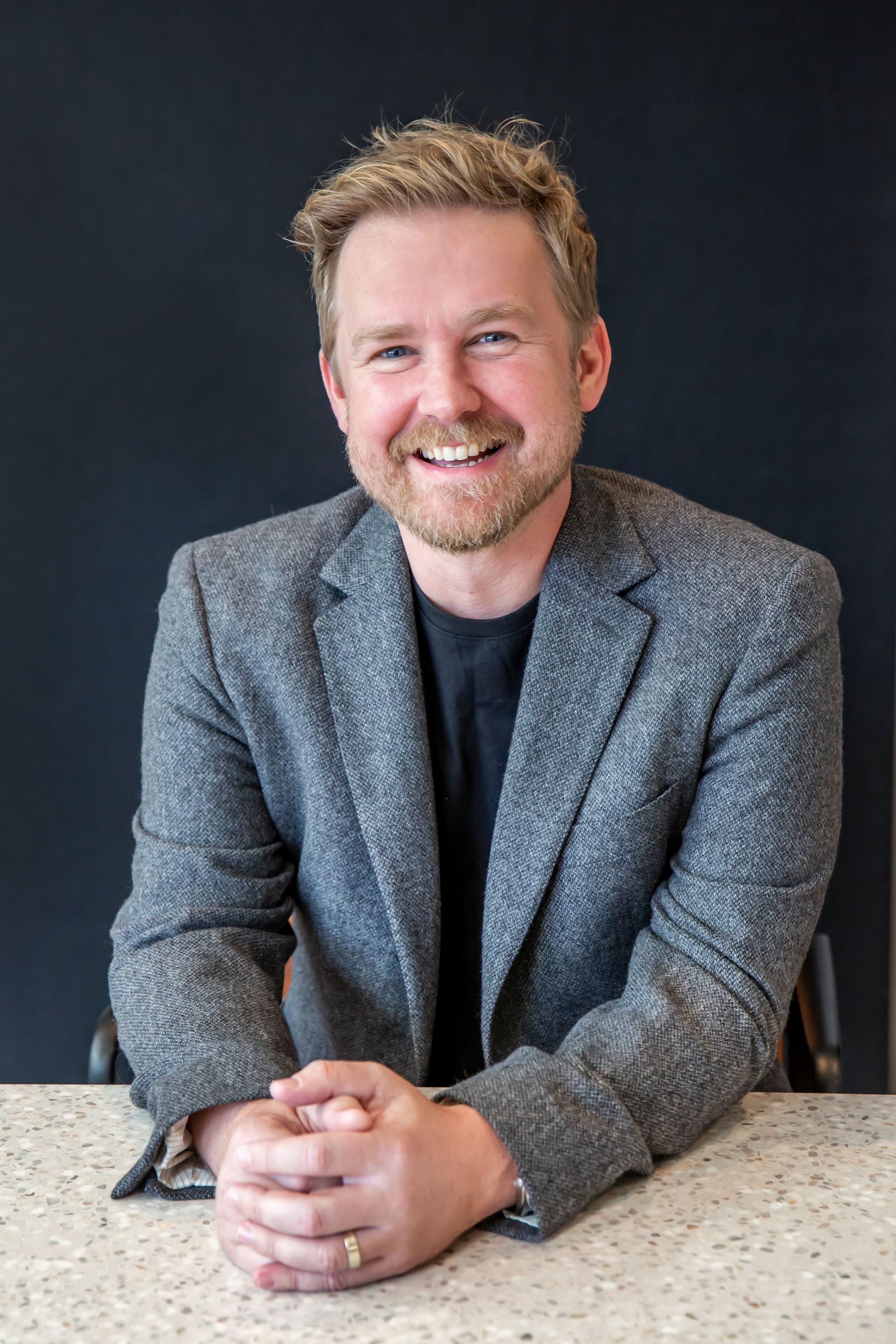 Smiling man with blonde hair in a gray blazer, leaning on a counter with hands clasped, against a dark wall.