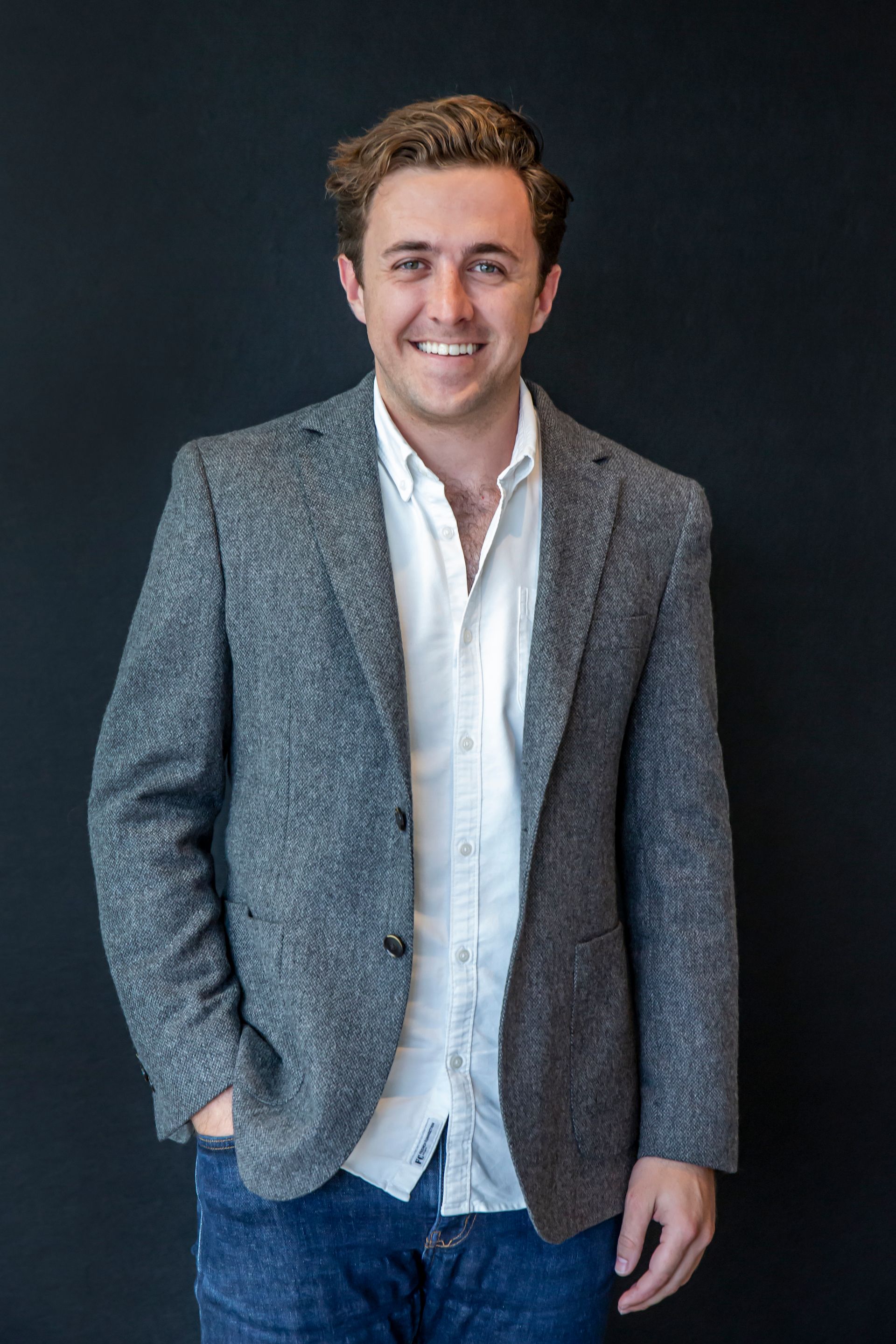 Man in gray blazer and white shirt, smiling, standing against a dark wall.