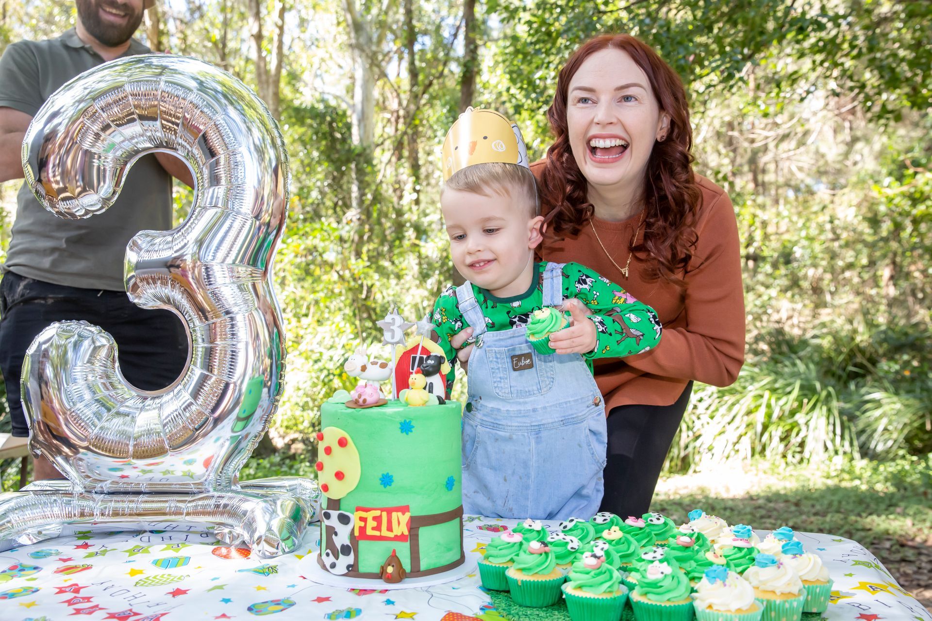 Child celebrating 3rd birthday, laughing with parents near a cake and cupcakes outdoors.