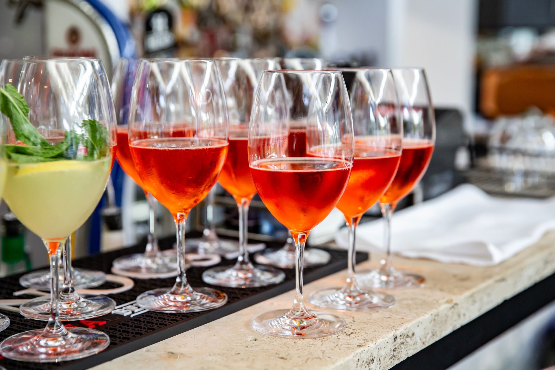 Row of Aperol Spritz cocktails, vibrant orange, in wine glasses on a bar. A single limoncello cocktail is on the left.