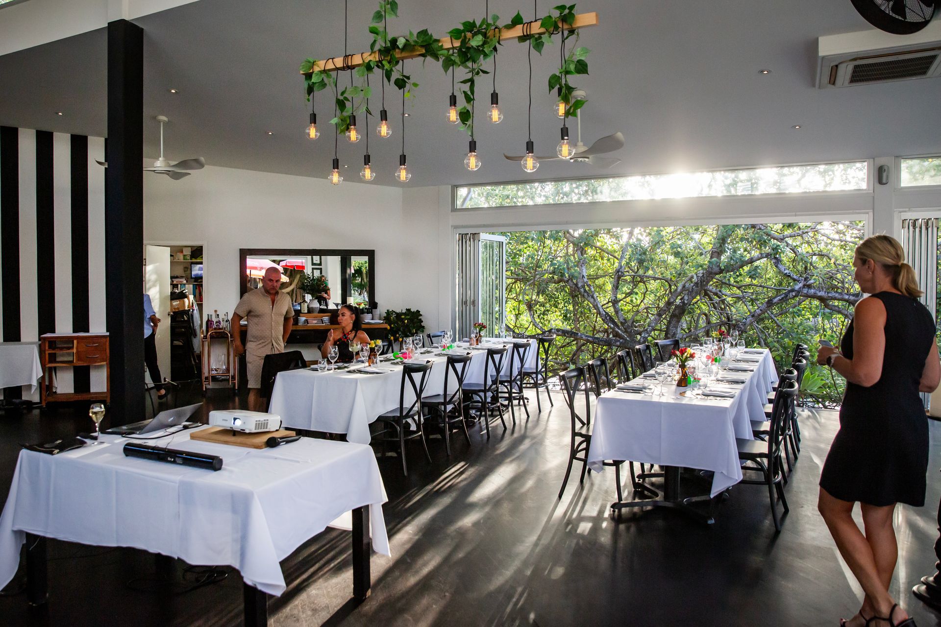 Bright dining room with tables set for a meal, a woman looks at the tables, sunlight streams through large windows.