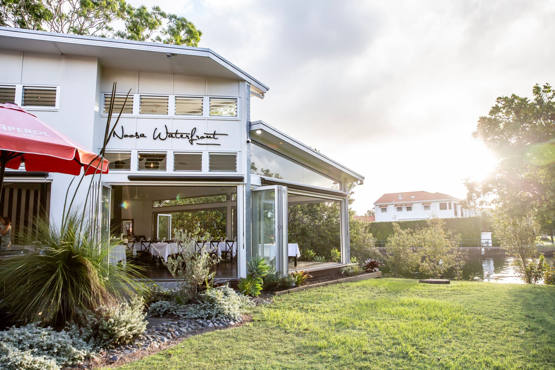 White waterfront restaurant with open doors, green lawn, trees, and bright sunlight.