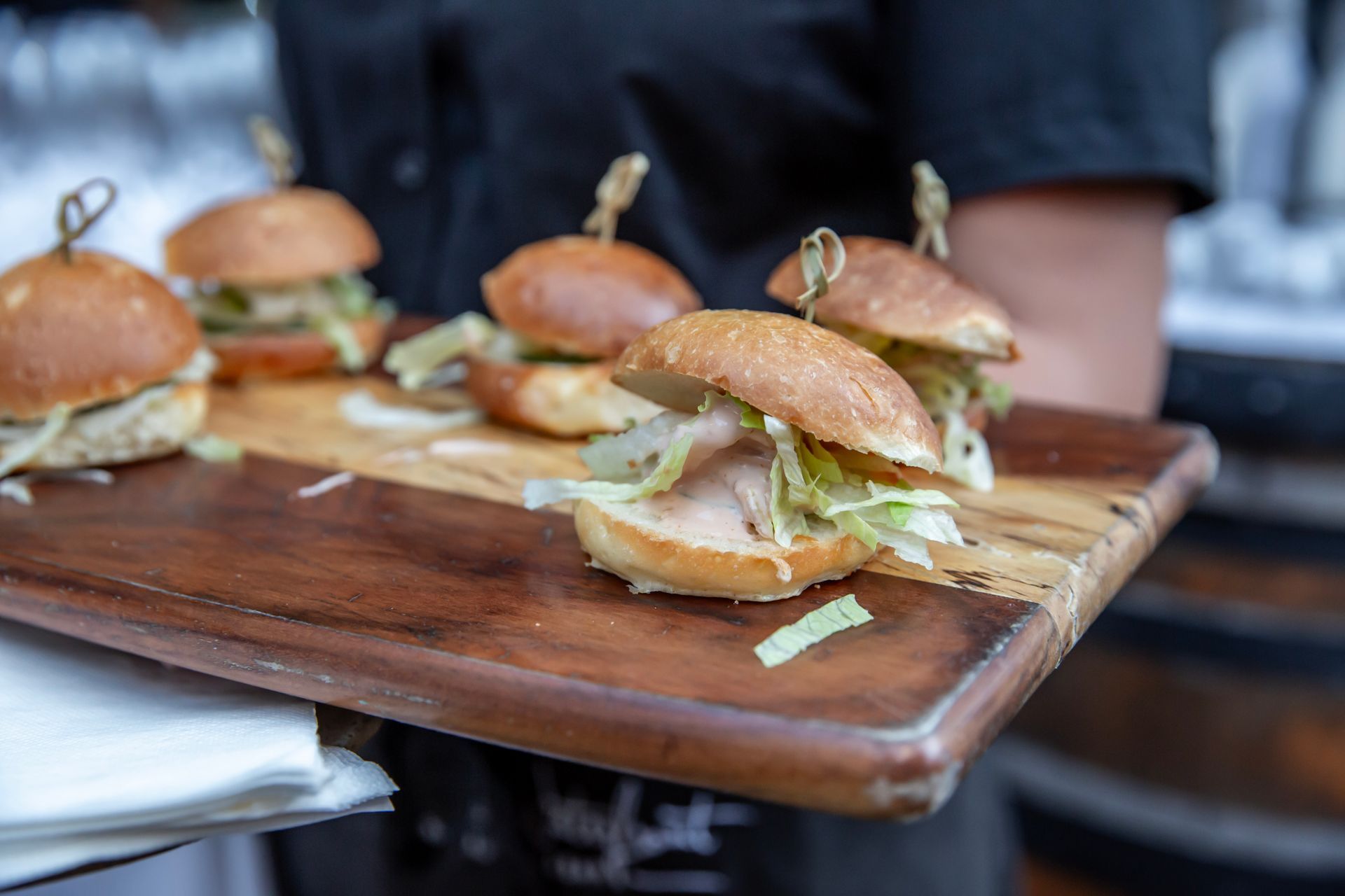 A server holding a wooden tray with six small sliders topped with lettuce and skewers.