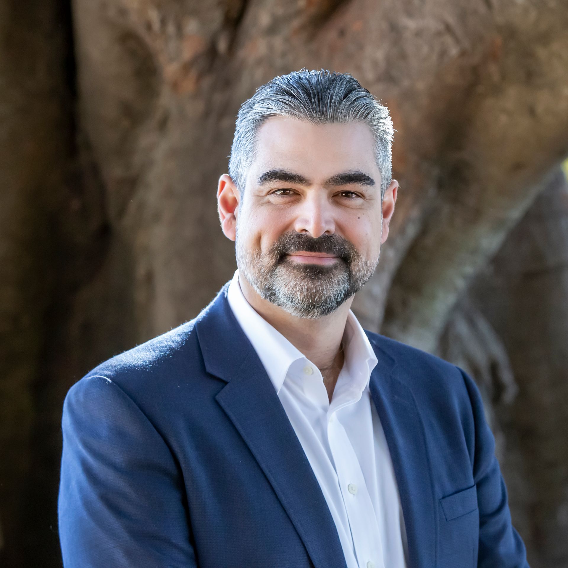 A man with graying hair and beard in a blue suit smiles in front of a tree.