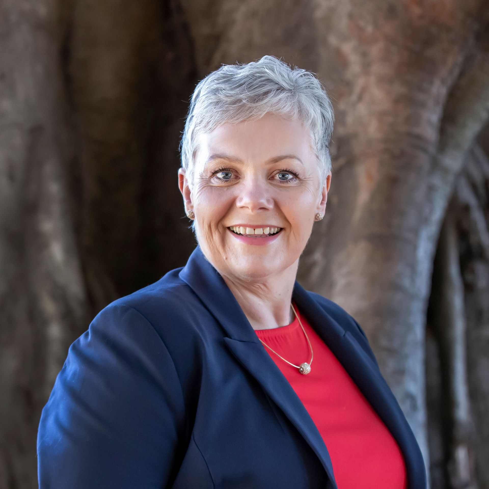 Woman with short gray hair, wearing a blue blazer and red top, smiling against a tree trunk.