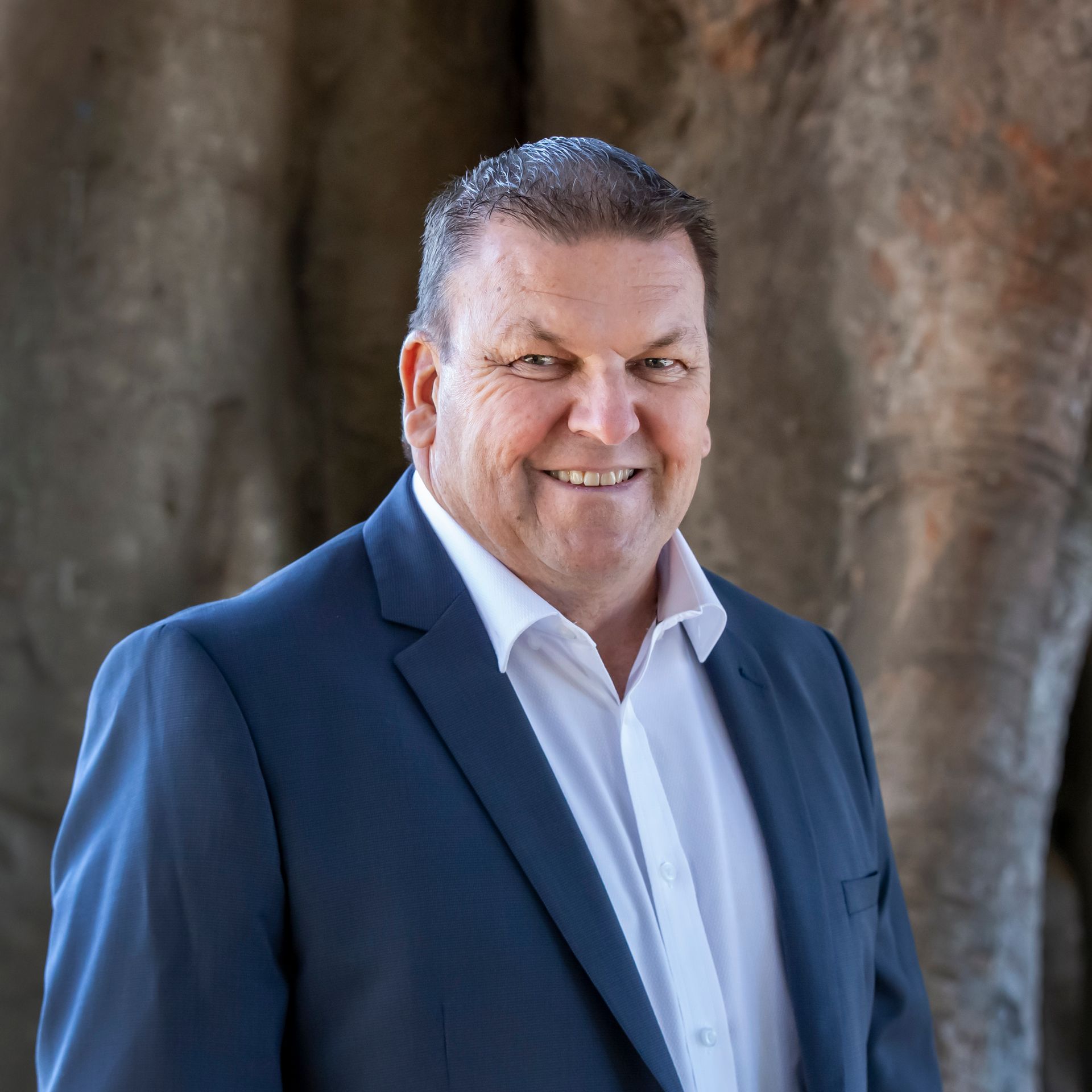 Man in suit smiling, standing in front of tree trunk.