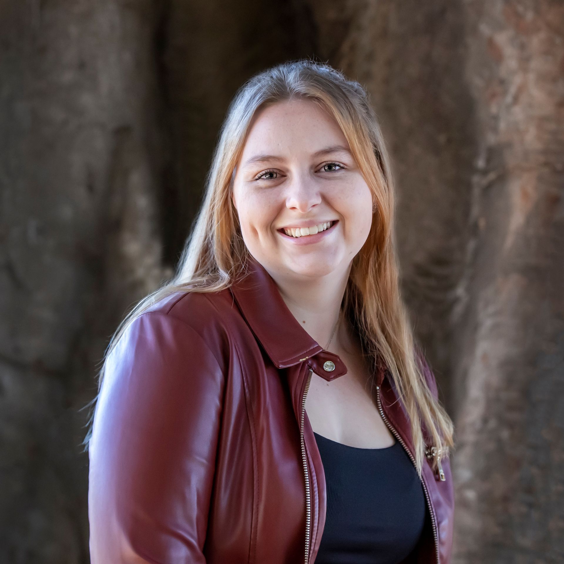 Woman with blonde hair, smiling in a maroon leather jacket and black top, set against a blurred brown background.