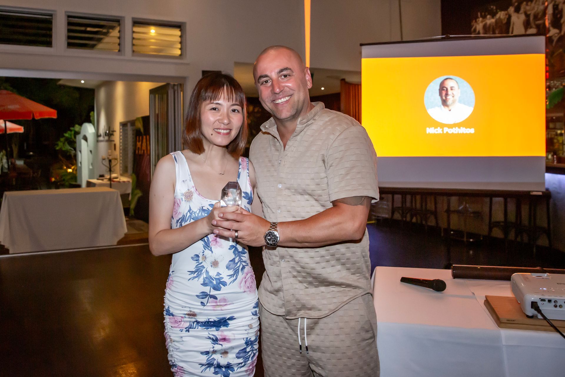 Woman in floral dress and man in tan suit pose, holding an award. Presentation screen in background.