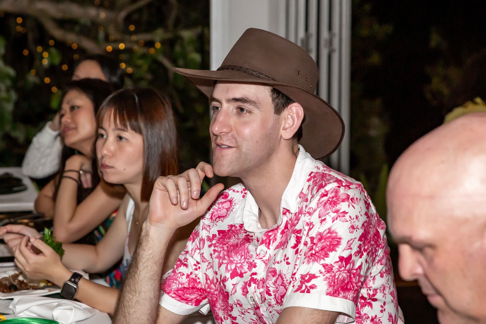 Man in cowboy hat at a gathering, wearing a pink and white floral shirt.