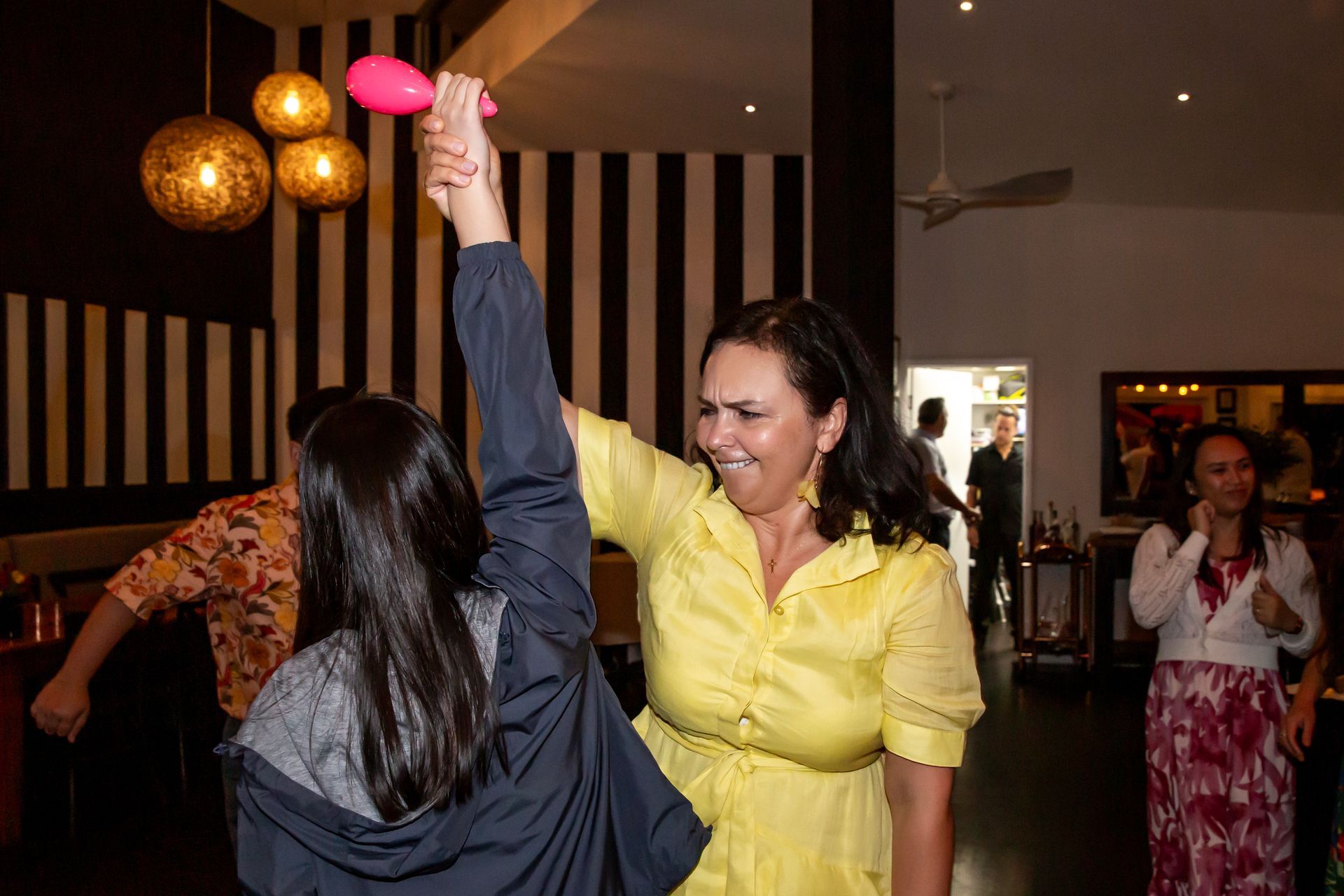 Woman in yellow dress dances, holding maraca, with another person in a room with others.
