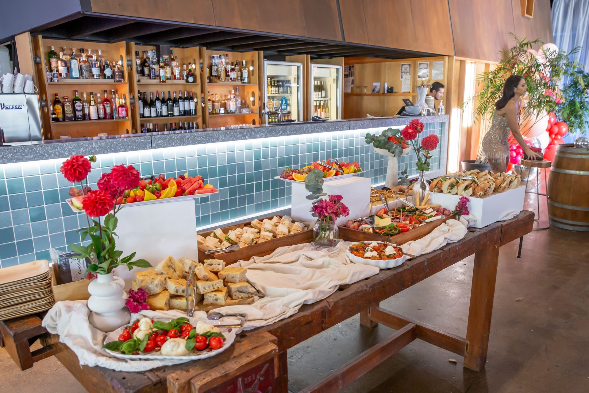 Buffet table with food and flowers set up in front of a bar; a person is standing nearby.