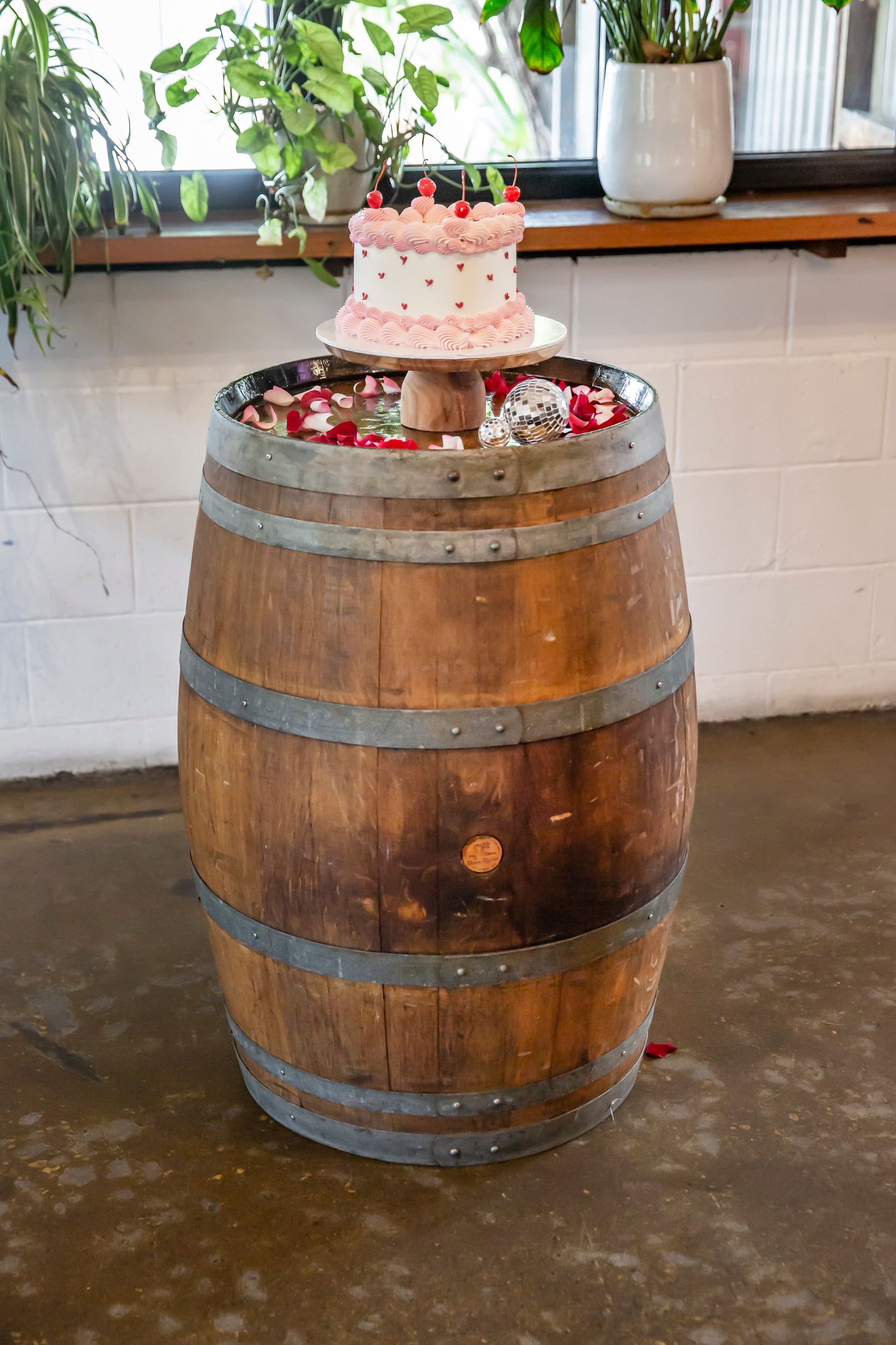Wedding cake on a wooden barrel surrounded by rose petals; set indoors with greenery and windows.
