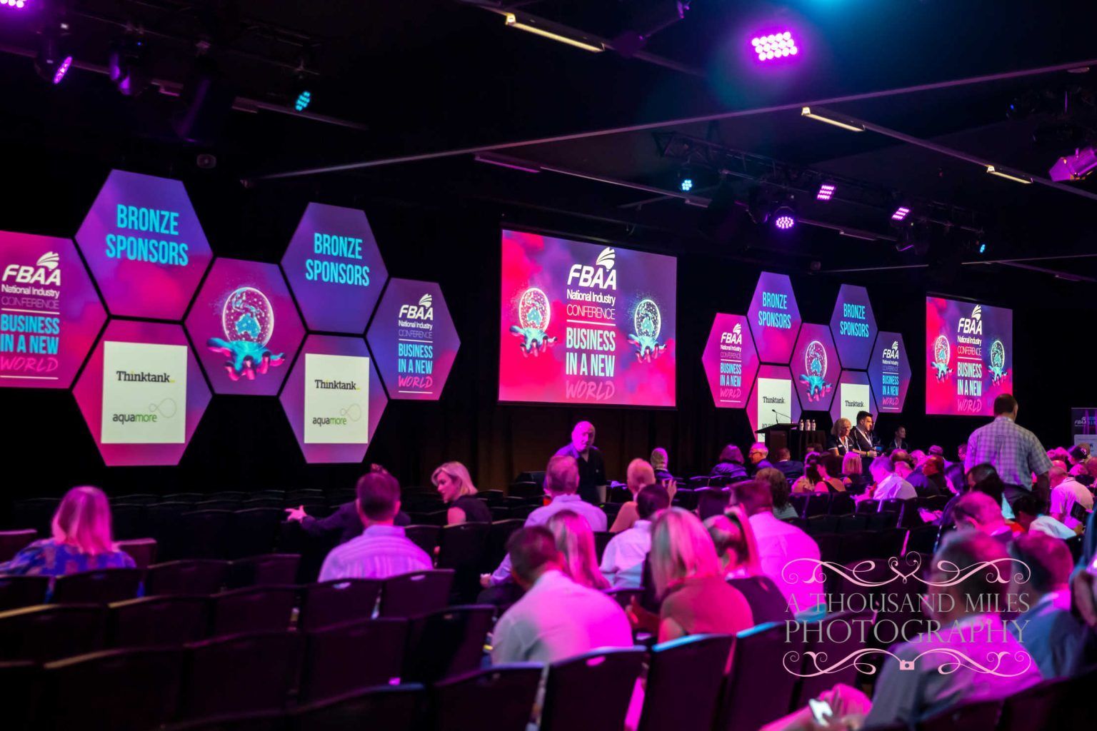 Audience at a conference, looking towards a stage with sponsor logos on screens, lit with pink and purple lights.