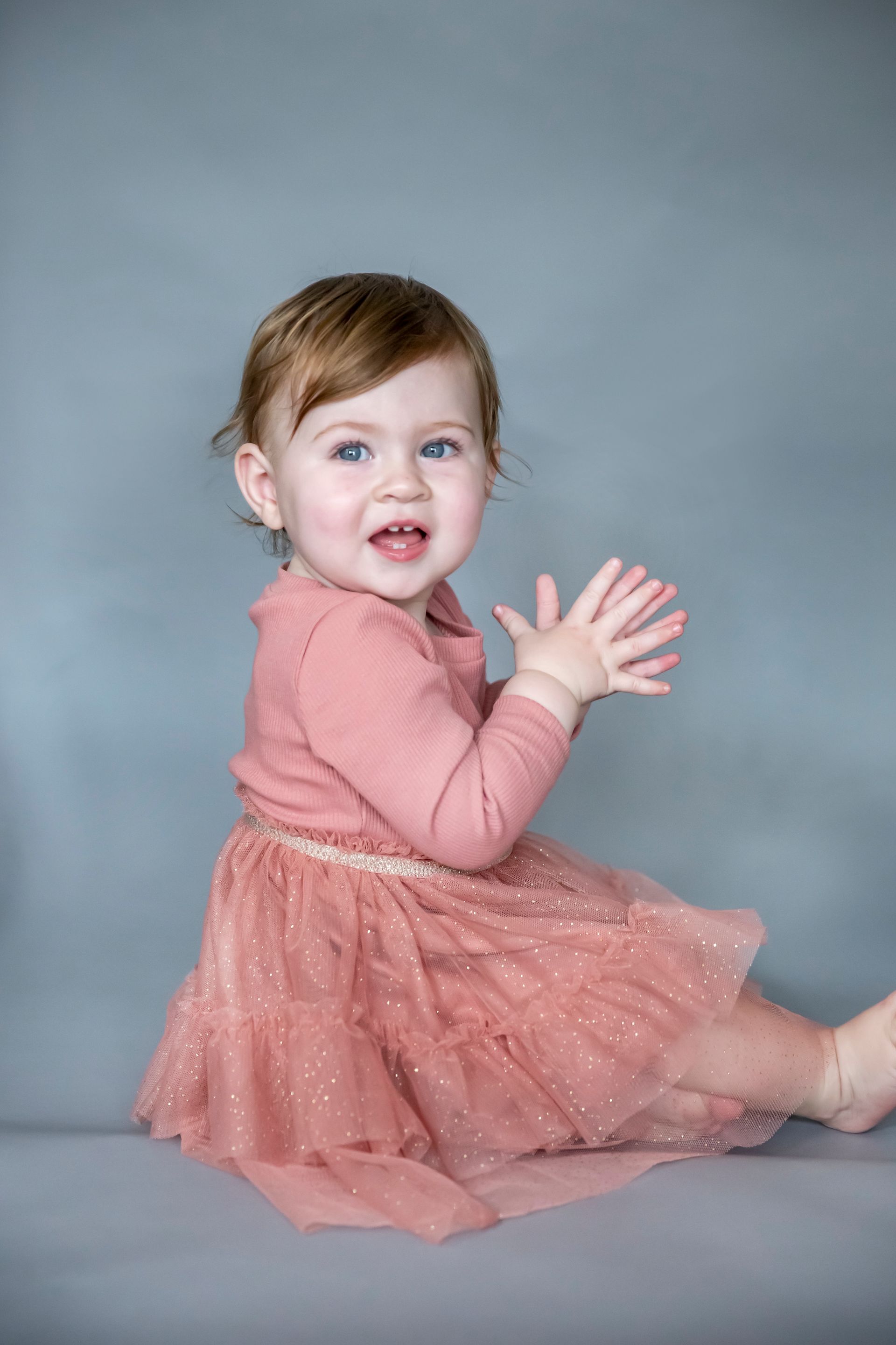 A toddler with rosy cheeks claps while wearing a pink sweater and skirt, seated against a gray backdrop.