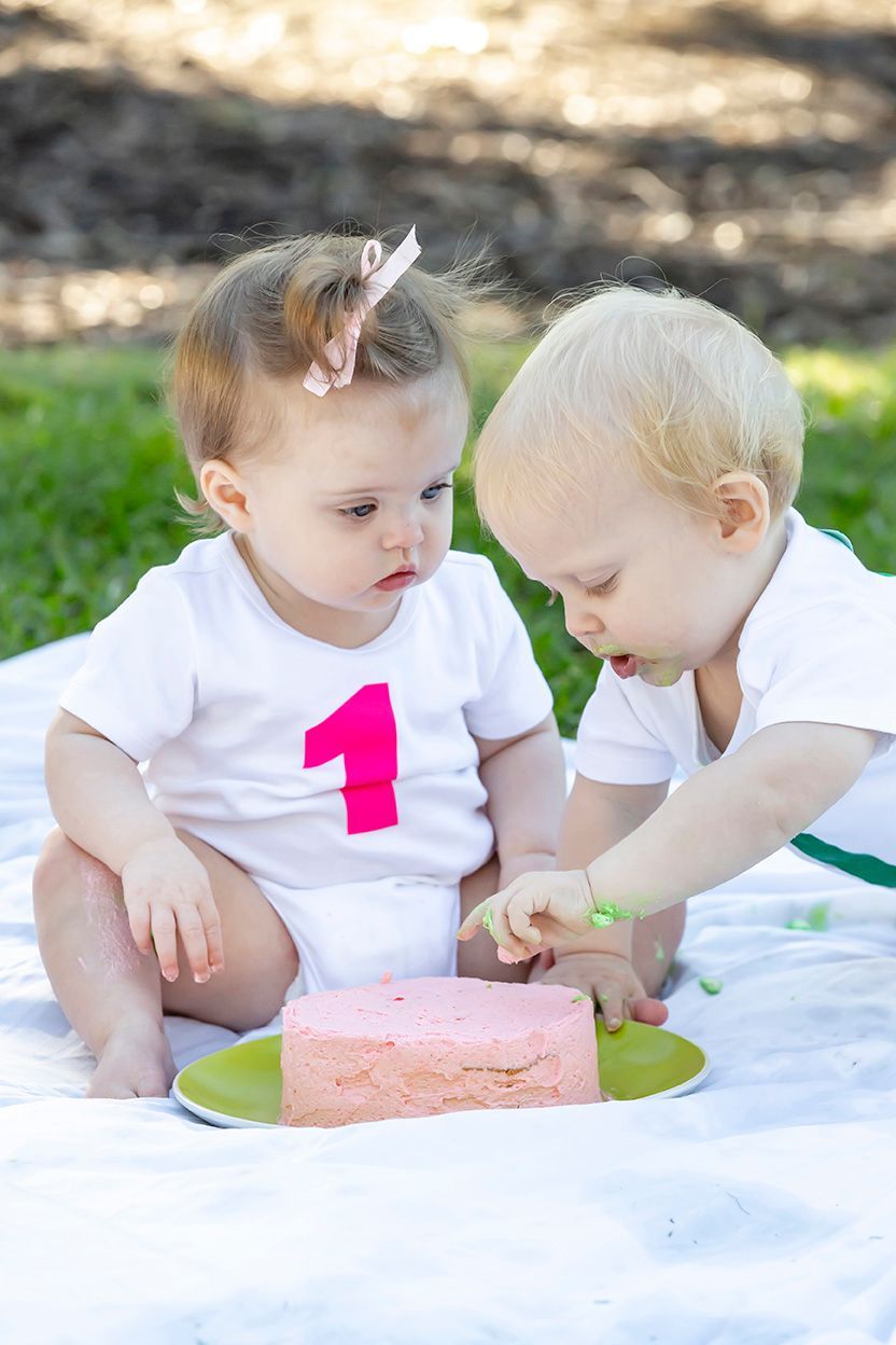 Two babies at a birthday celebration; one in a white onesie with pink 