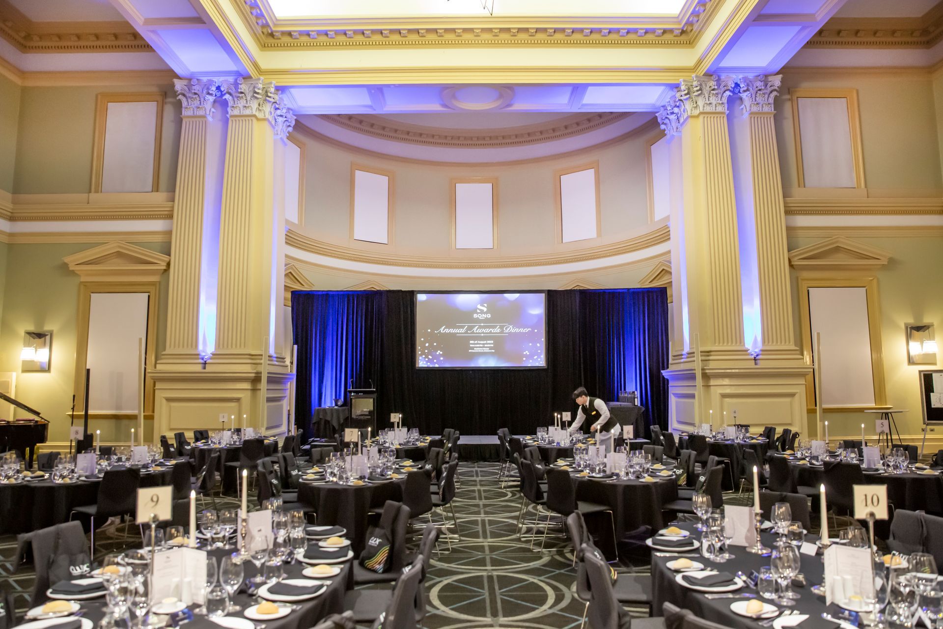 Elegant banquet hall with tables set for a formal event. Stage, columns, and blue and gold lighting.