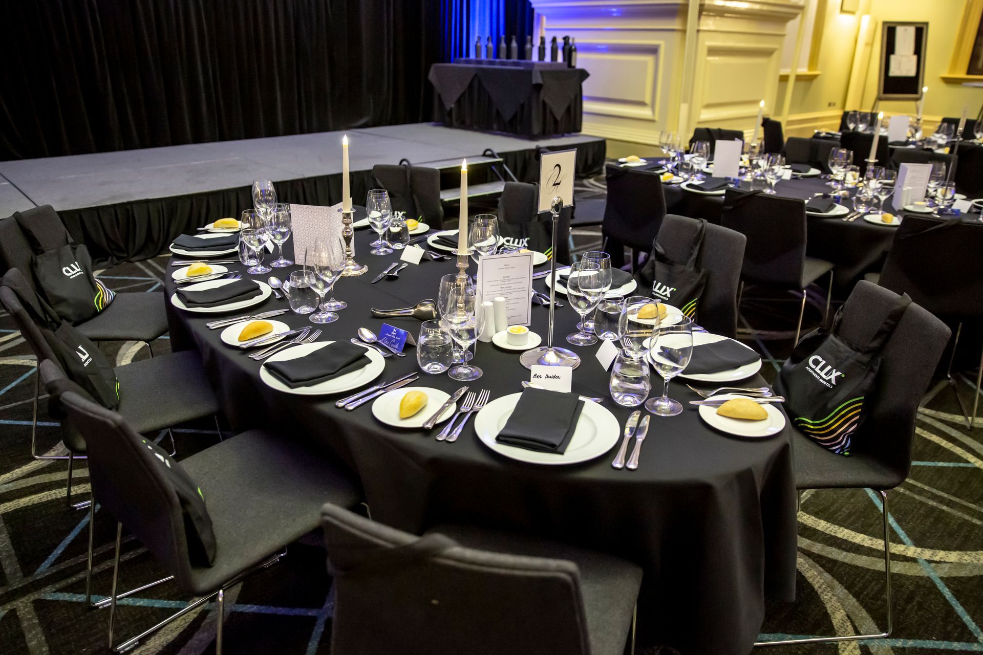 Oval tables set for a formal dinner, black tablecloths, white plates, and silverware, in a banquet hall.