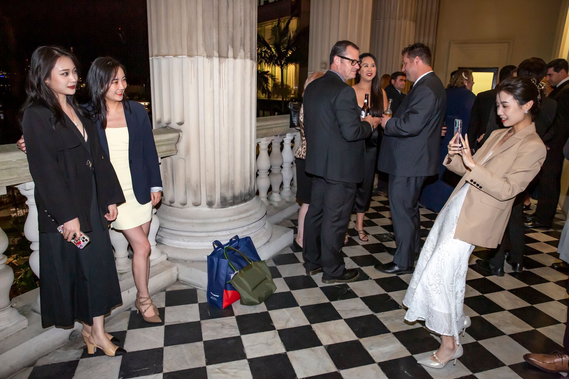 People at an outdoor event. Woman takes photo of two others. Black and white tiled floor. Evening setting.